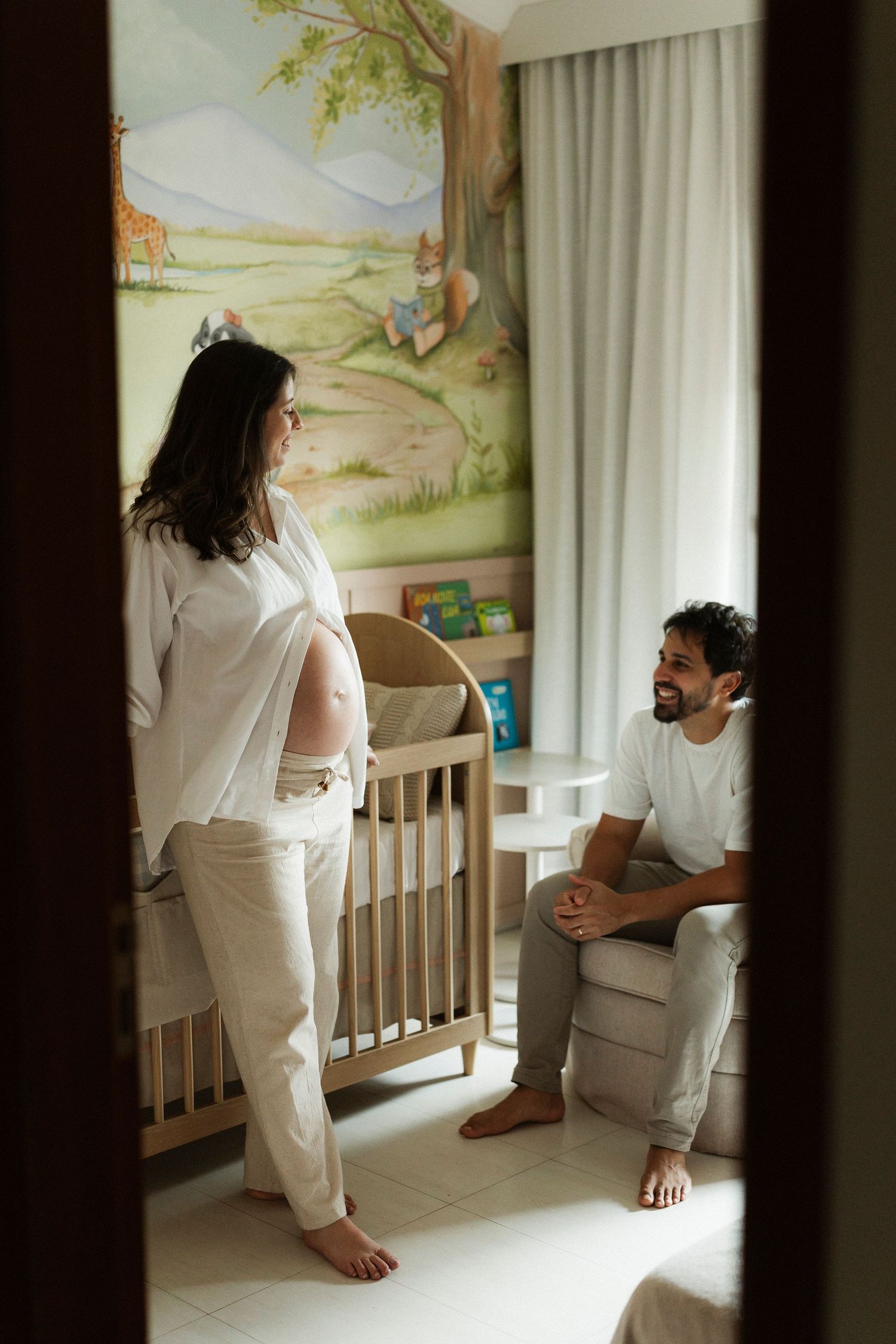 Pregnant woman and smiling man share a tender moment in a beautifully decorated nursery room with nature mural.