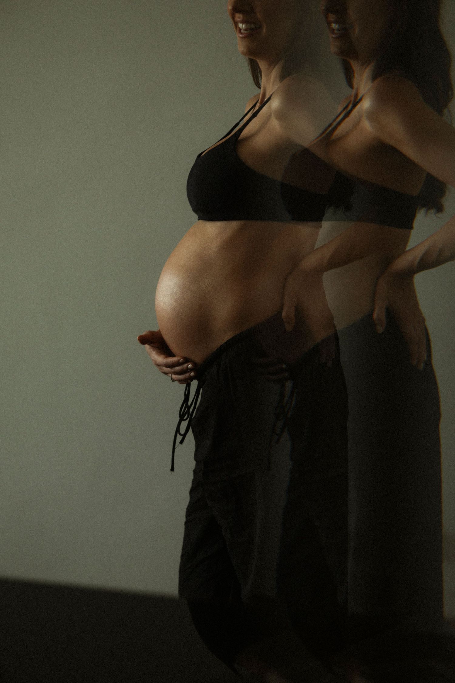 Artistic silhouette of pregnant woman in black sports bra and pants with reflection against moody lighting.
