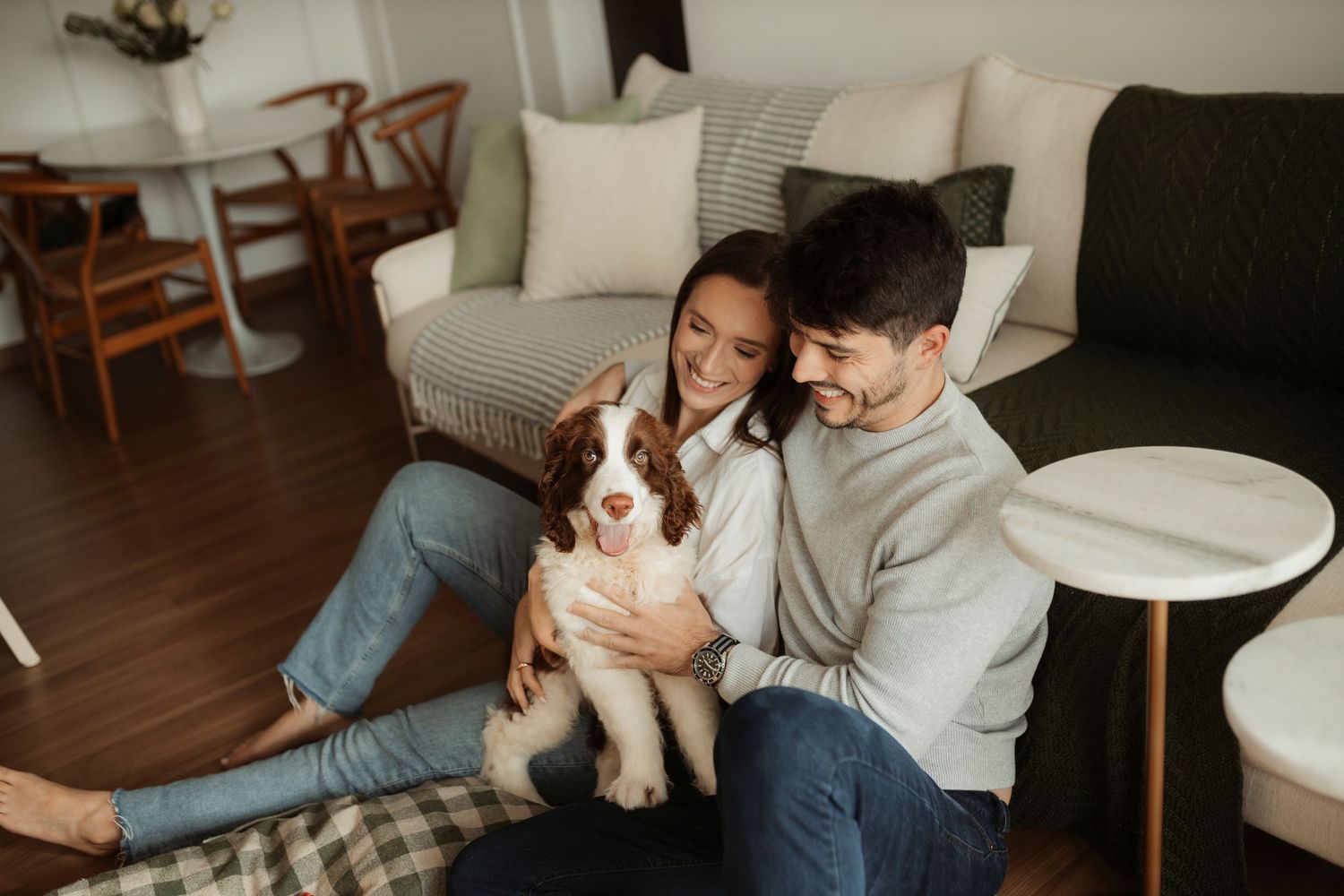 A couple relaxes on a couch with their small white dog in a cozy living room setting.