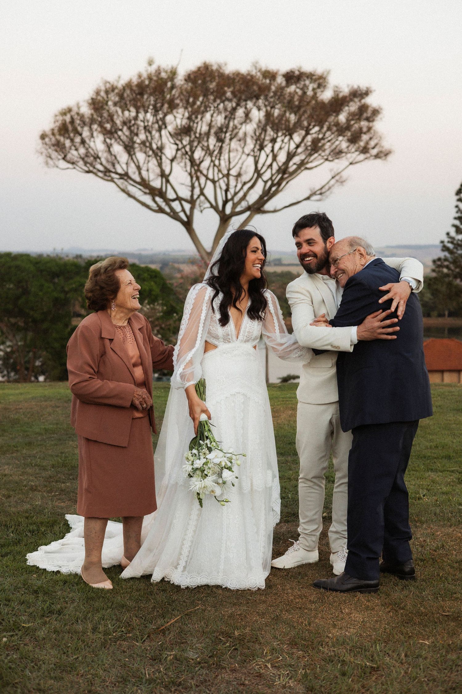 Wedding party gathers on hillside for candid group photos at sunset.