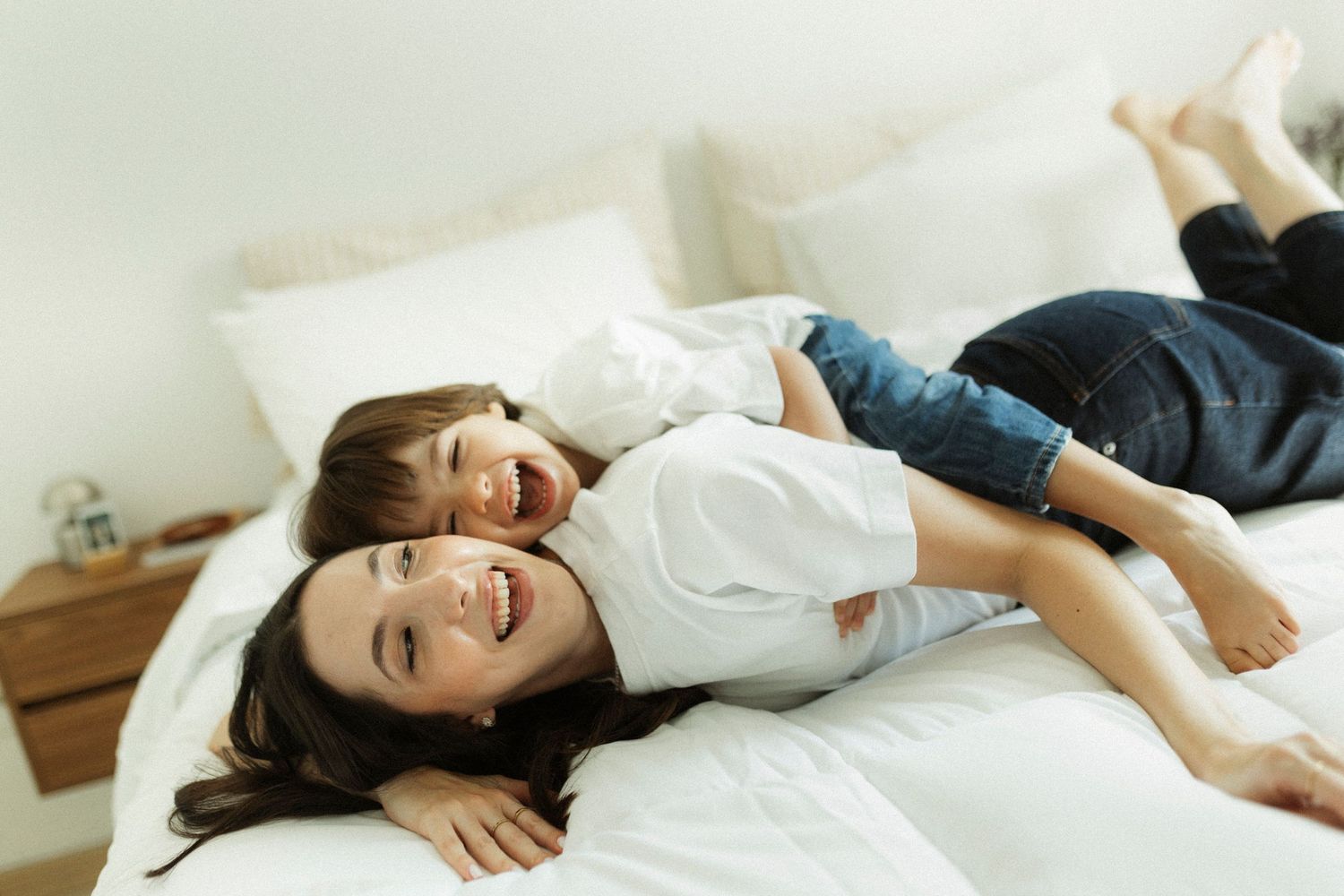 A joyful mother and young child laughing together while lying on a white bed, wearing casual white tops and jeans.