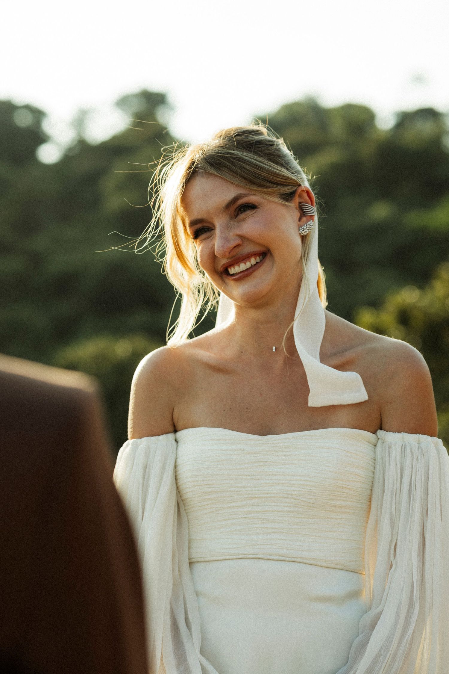 Smiling bride in white off-shoulder wedding dress outdoors with lush green trees in background.