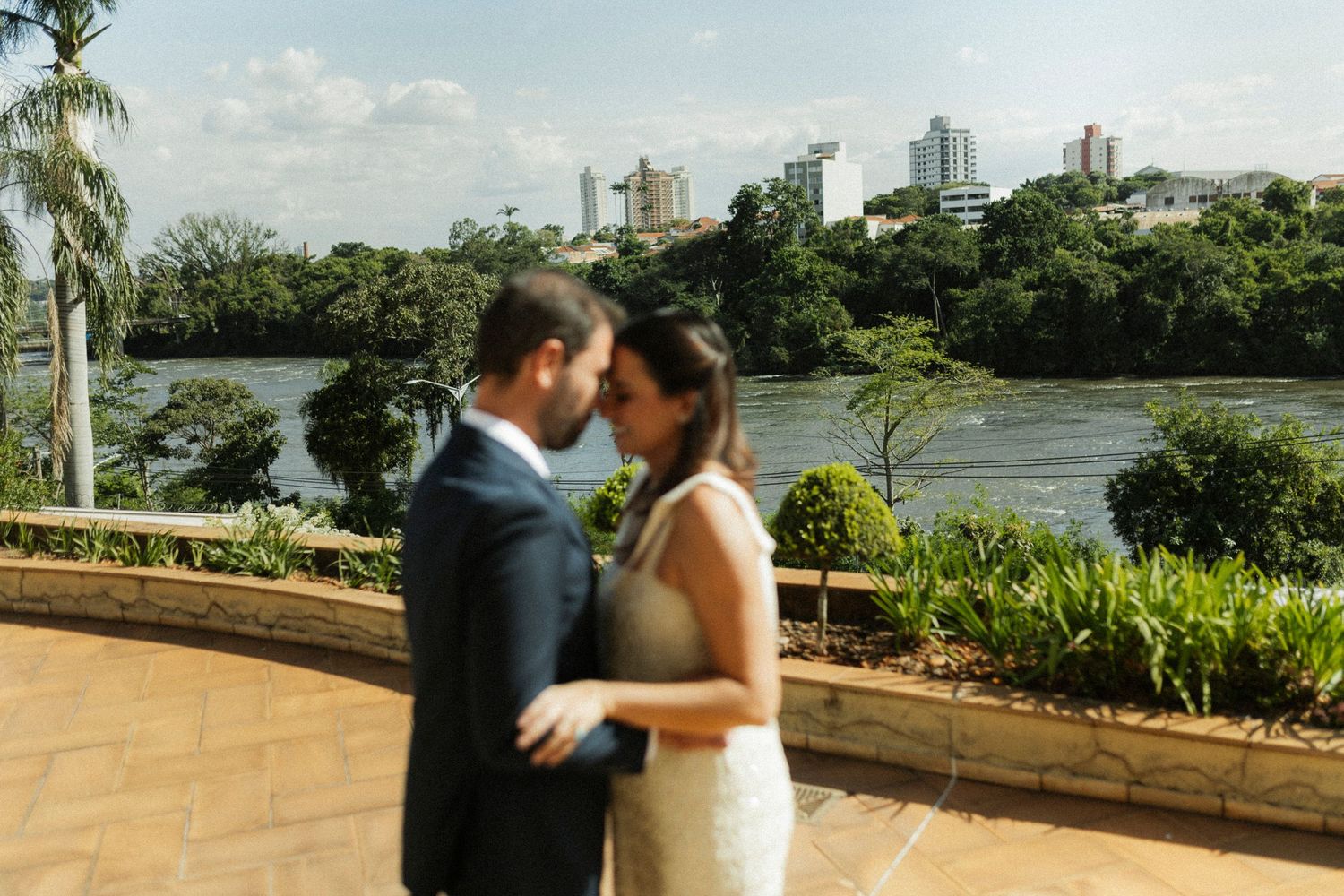 A couple shares a romantic moment on a terrace overlooking a river and city skyline, dressed in wedding attire.