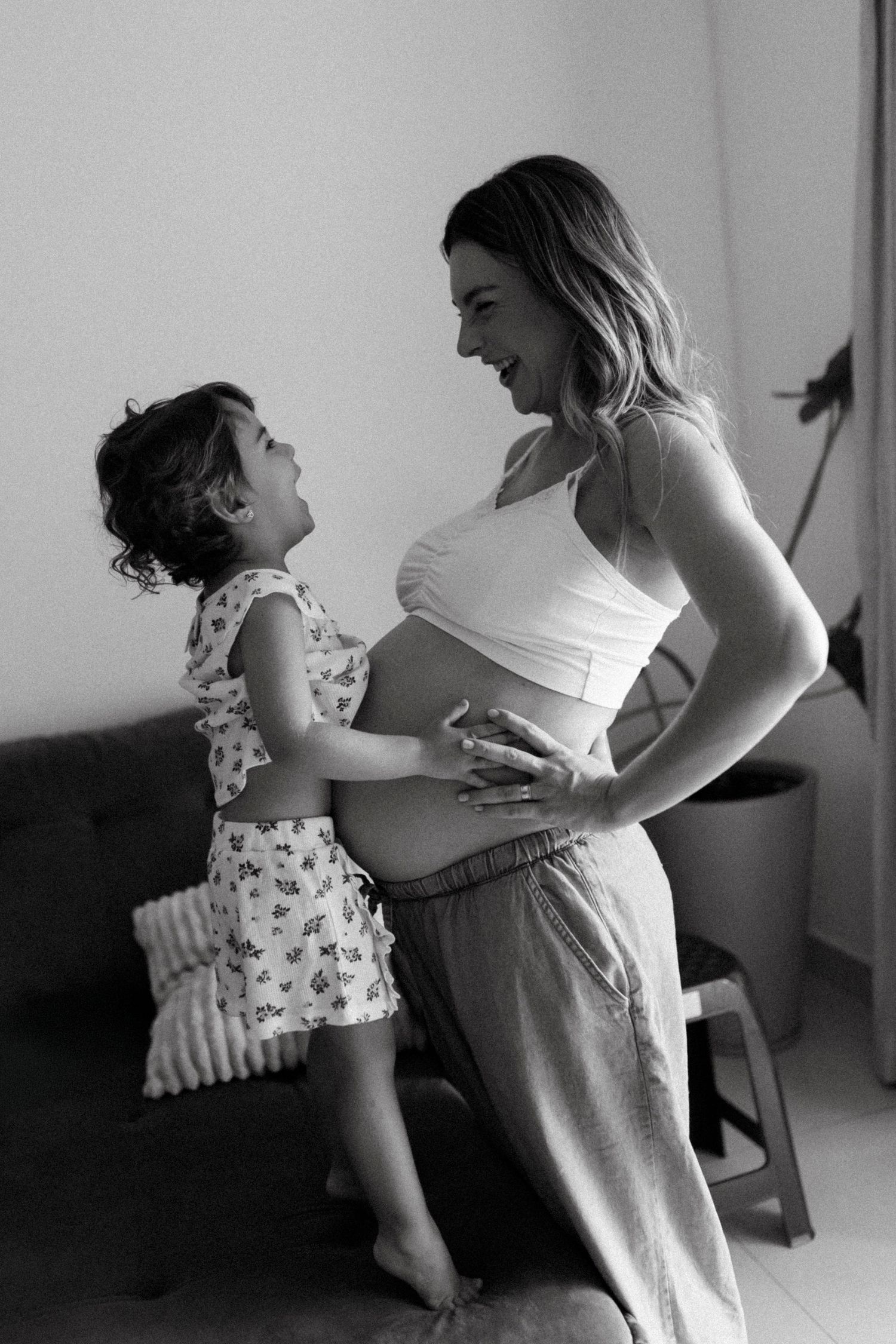 Black and white photo of a pregnant woman laughing with her young daughter touching her baby bump indoors.