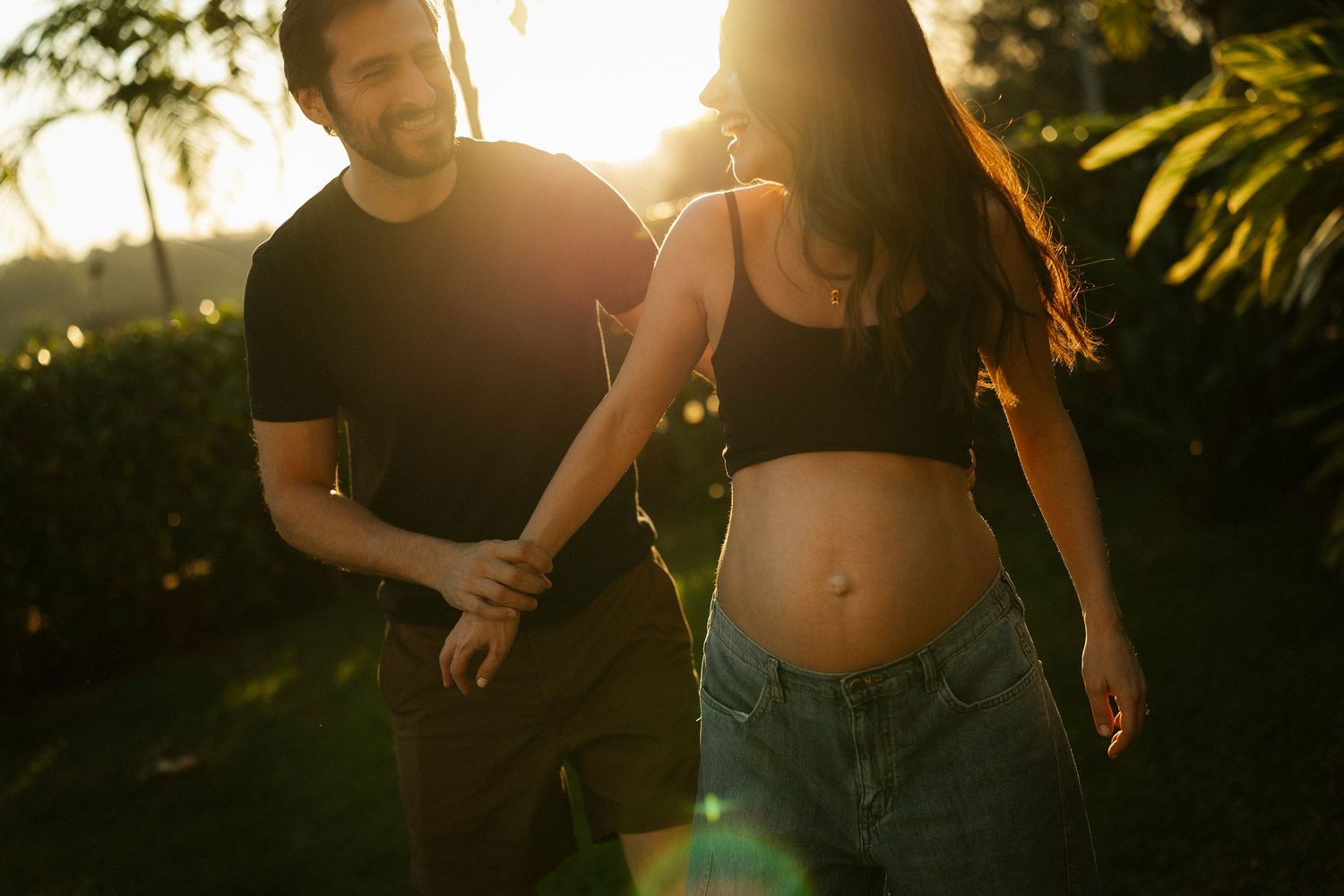Pregnant couple holding hands outdoors at golden hour, woman showing baby bump in crop top and jeans.