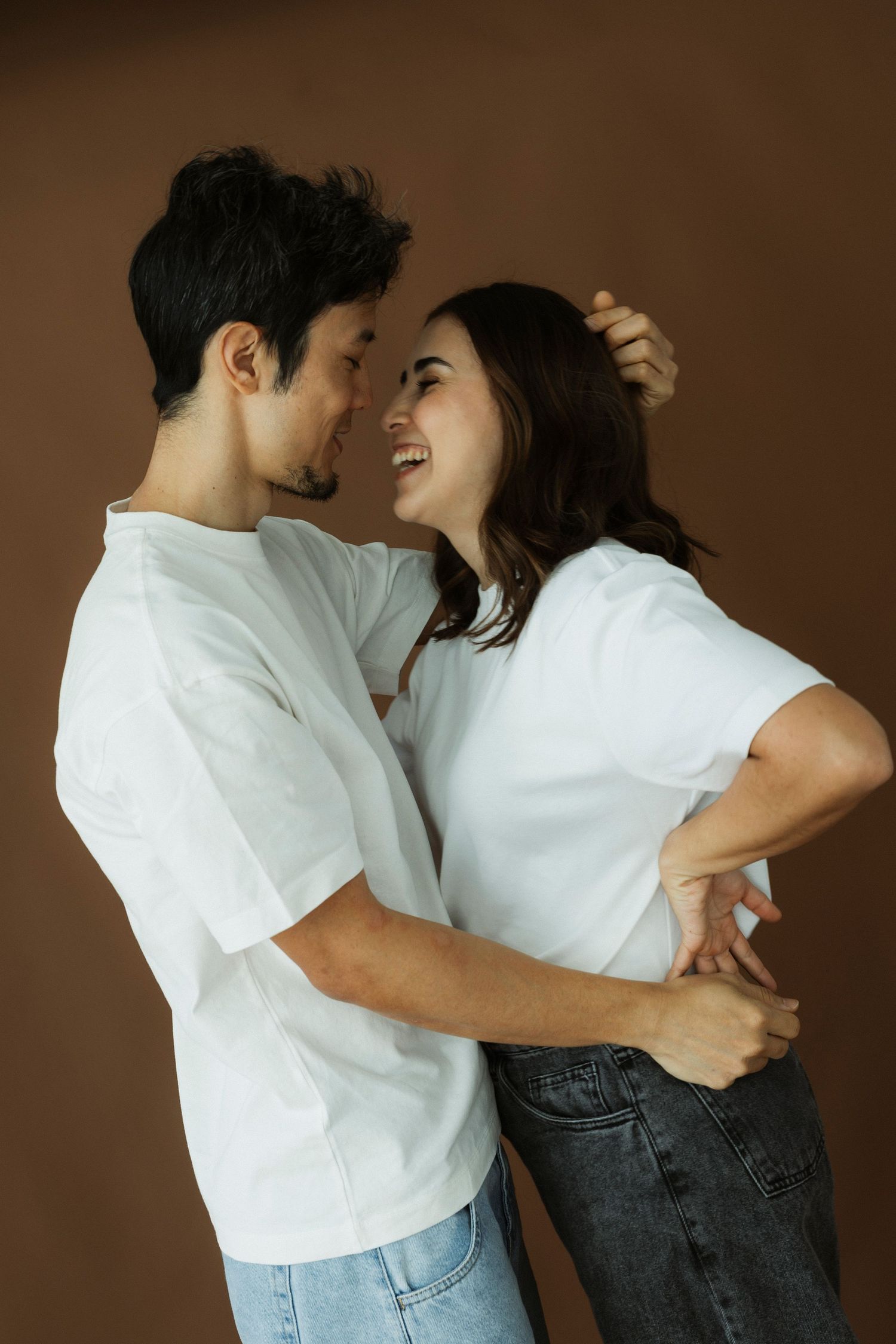 Couple in matching white t-shirts and jeans embracing and laughing together against a brown background.