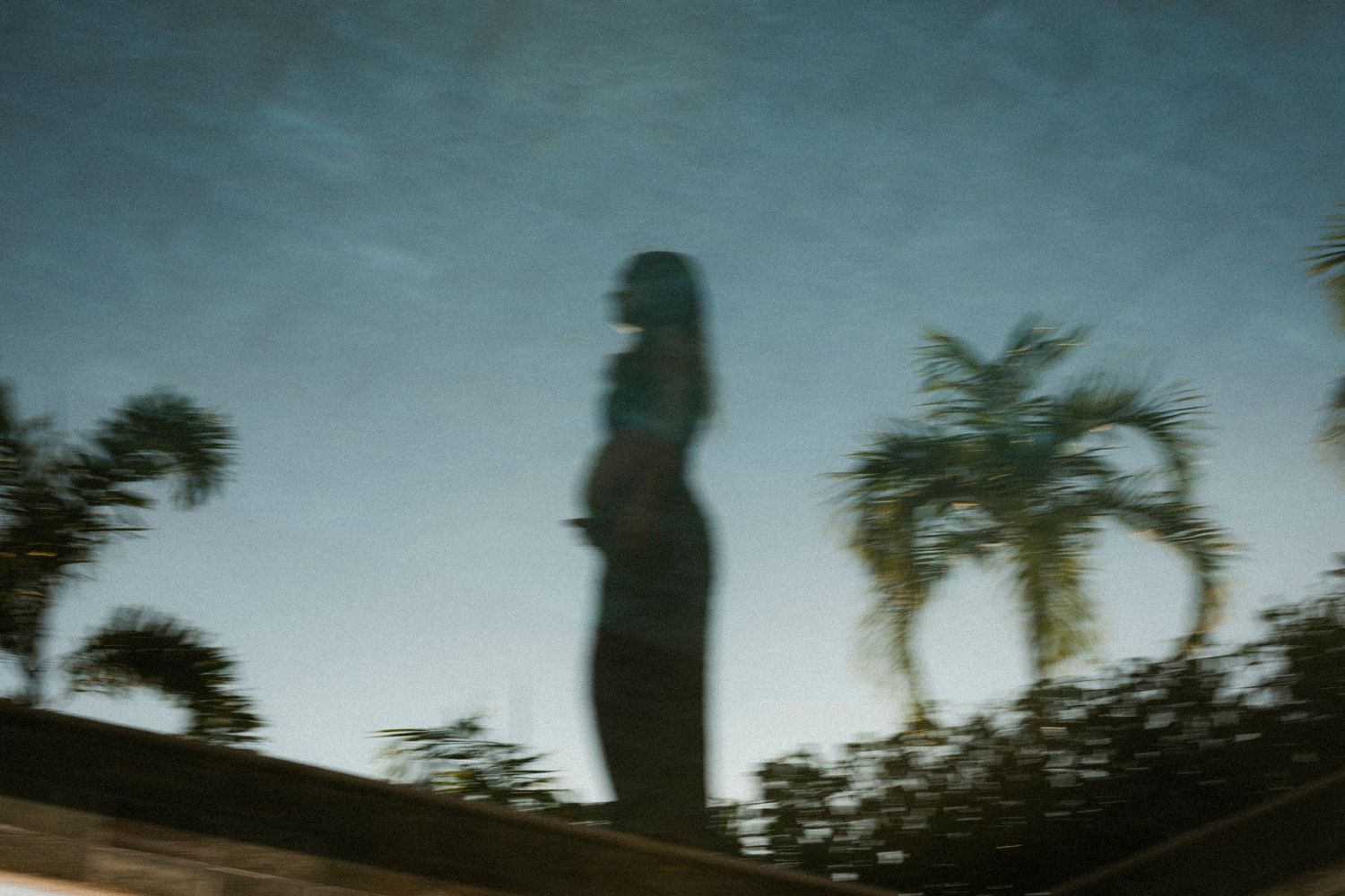 Blurry reflection of a person silhouette and palm trees in water under a blue sky.