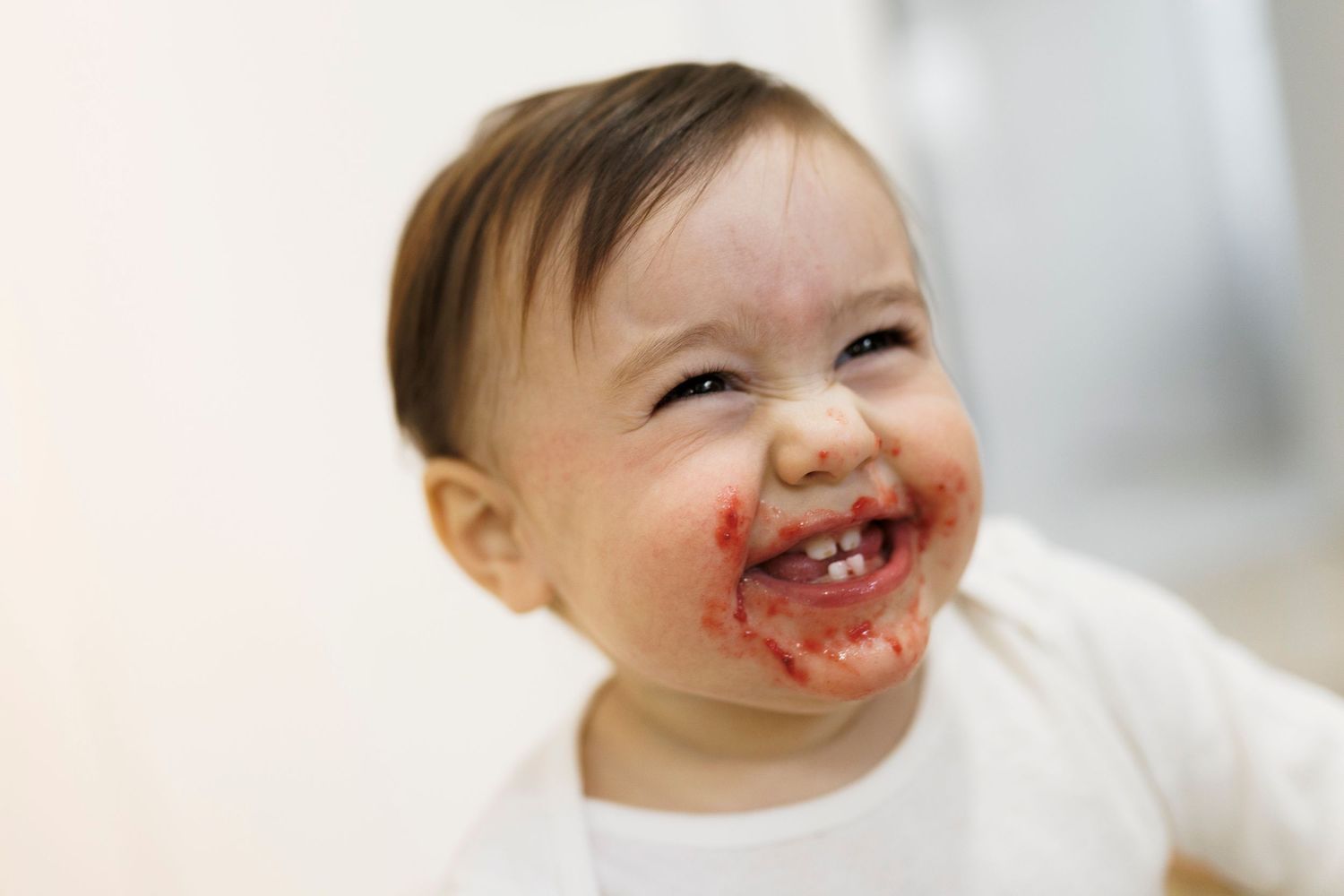 Baby with red fruit stains around mouth smiles joyfully during mealtime.
