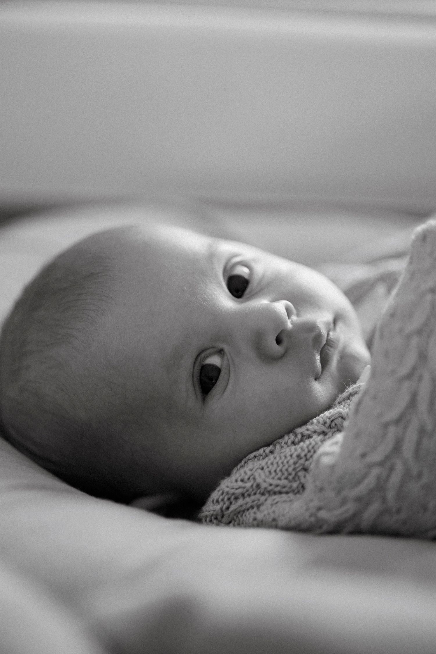 Black and white portrait of a newborn baby lying on a soft blanket, gazing upward with wide curious eyes.