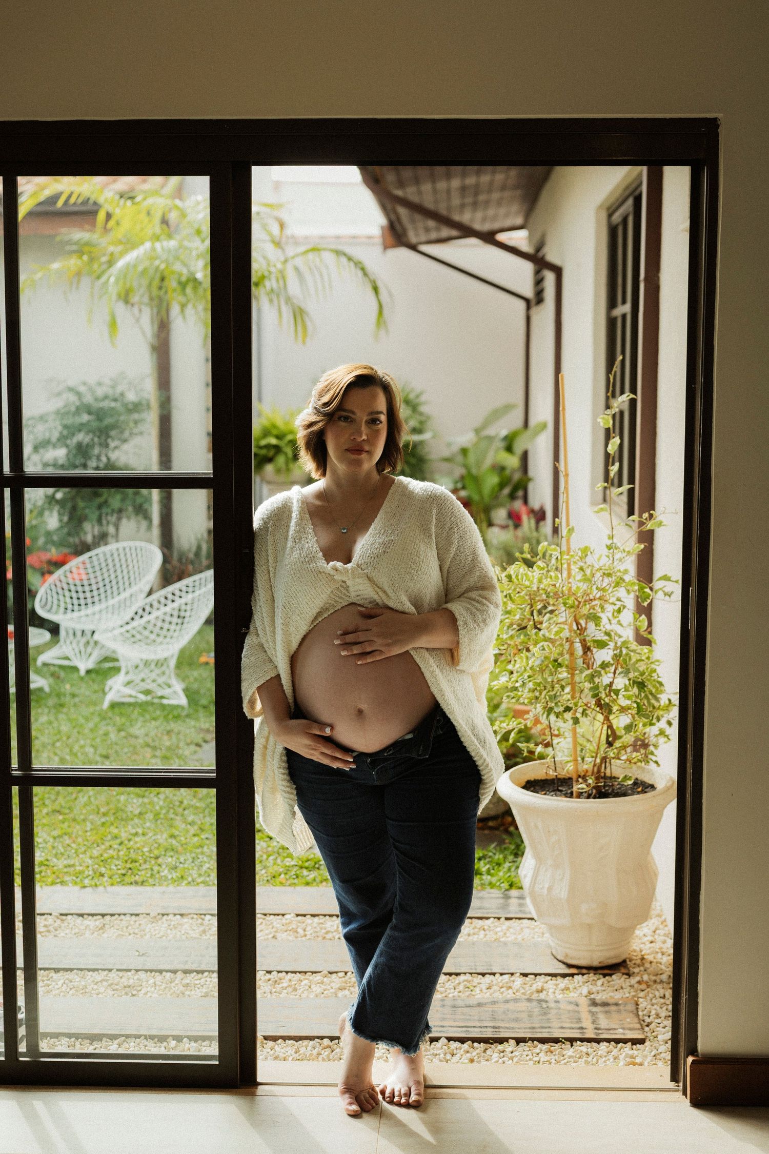 Pregnant woman in white cardigan and jeans cradling baby bump while standing in sunlit doorway with garden view.