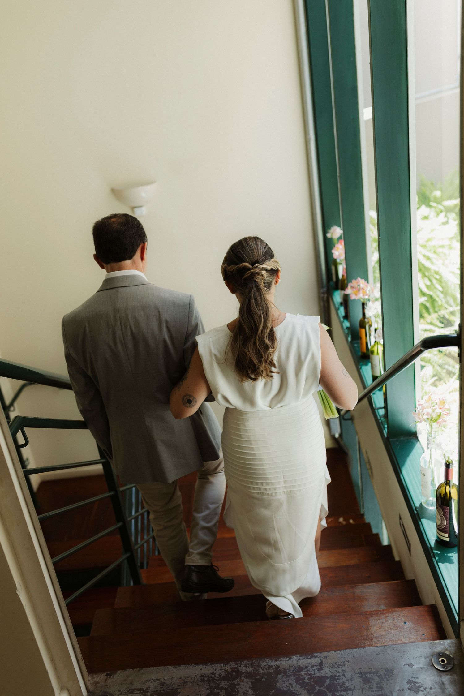 Couple in wedding attire walking up wooden stairs with green-trimmed windows.