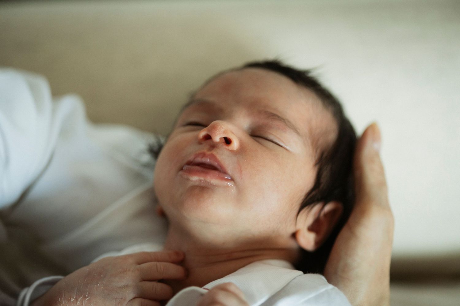 Peaceful newborn baby sleeping in parent's hands, wearing white onesie, with dark hair and serene expression.