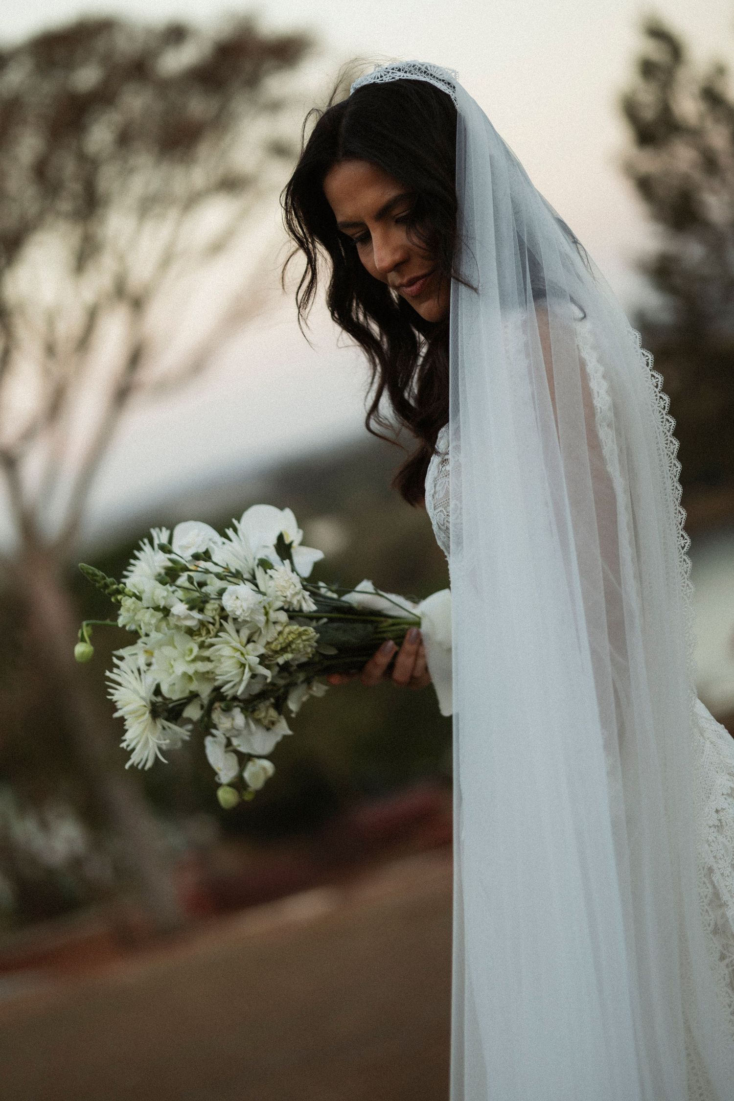 Bride in lace dress with flowing veil holds white bouquet during outdoor sunset portrait session.