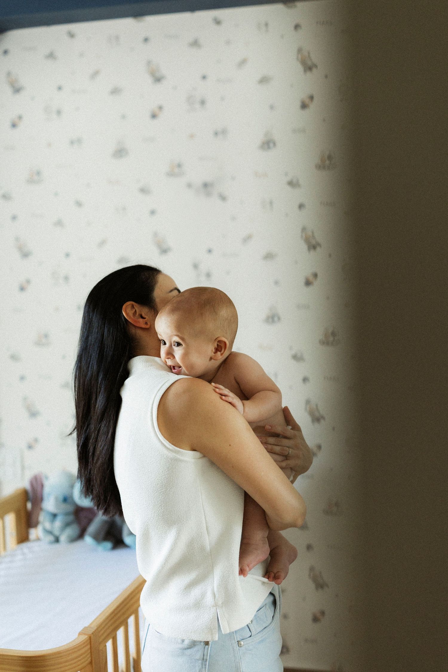A dark-haired mother in white top holds her smiling baby in a nursery with patterned wallpaper.