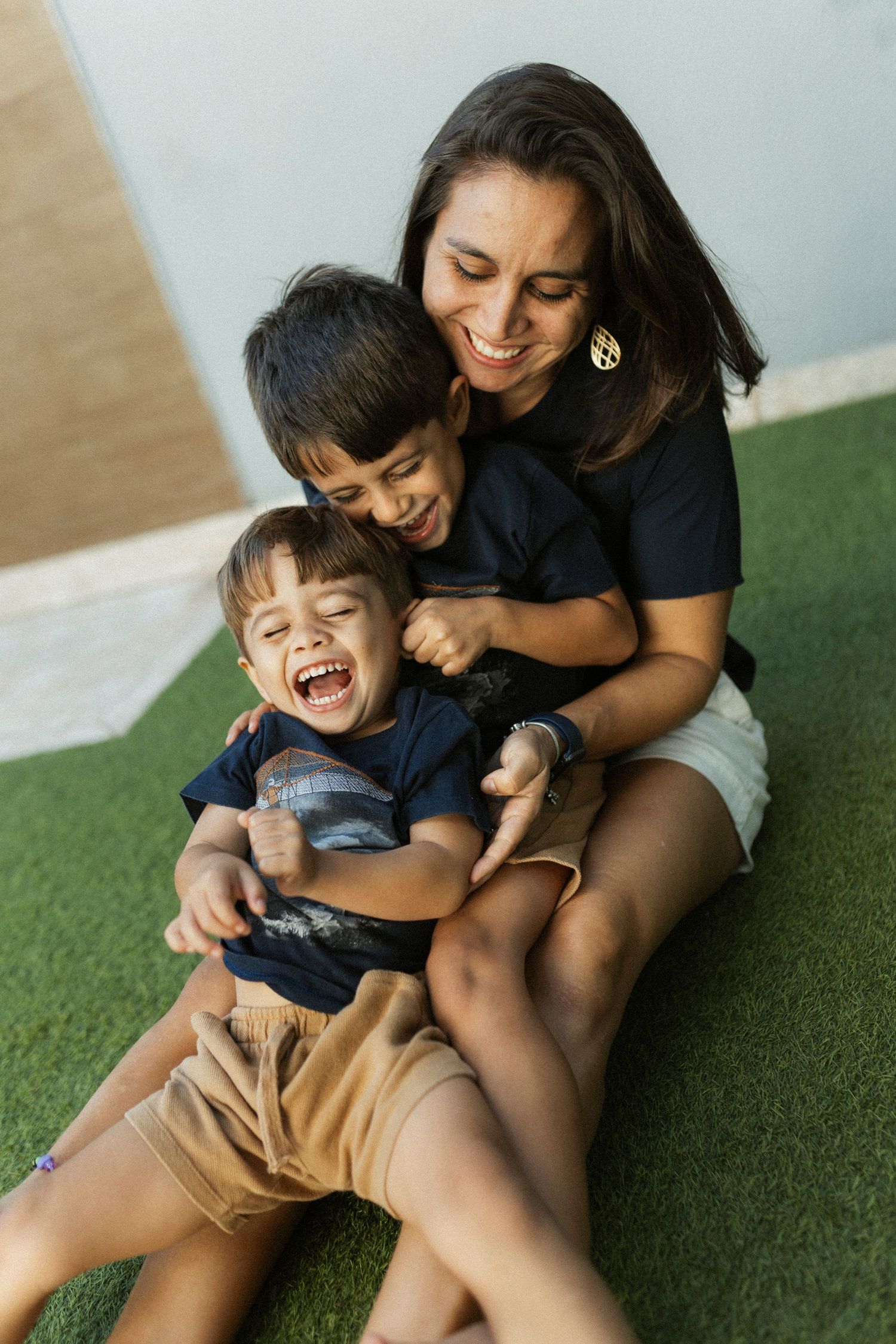 A smiling mother sits on grass hugging her two laughing young sons in matching navy shirts.
