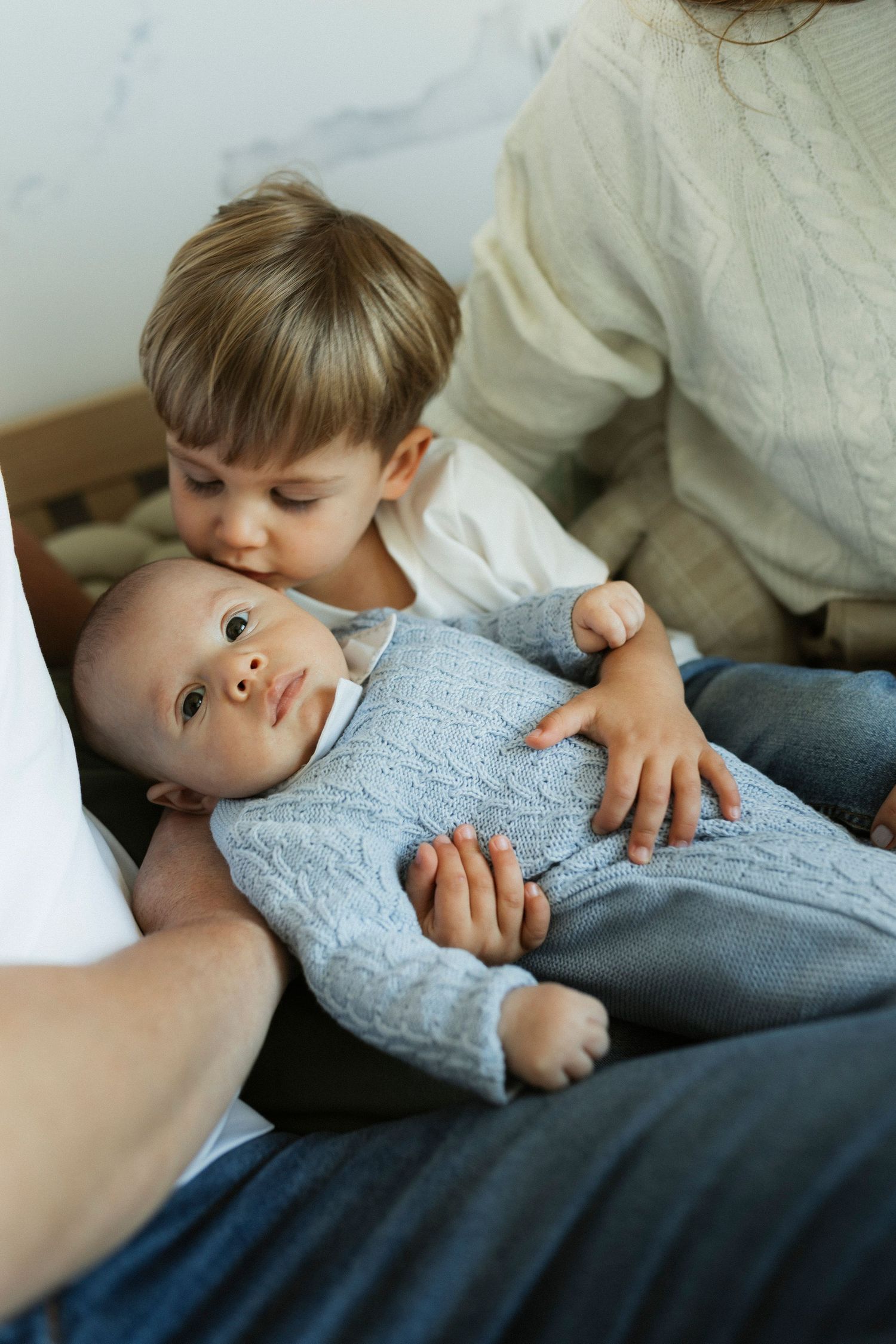 A young child gently kisses a newborn baby dressed in a blue knit onesie, held by a parent.