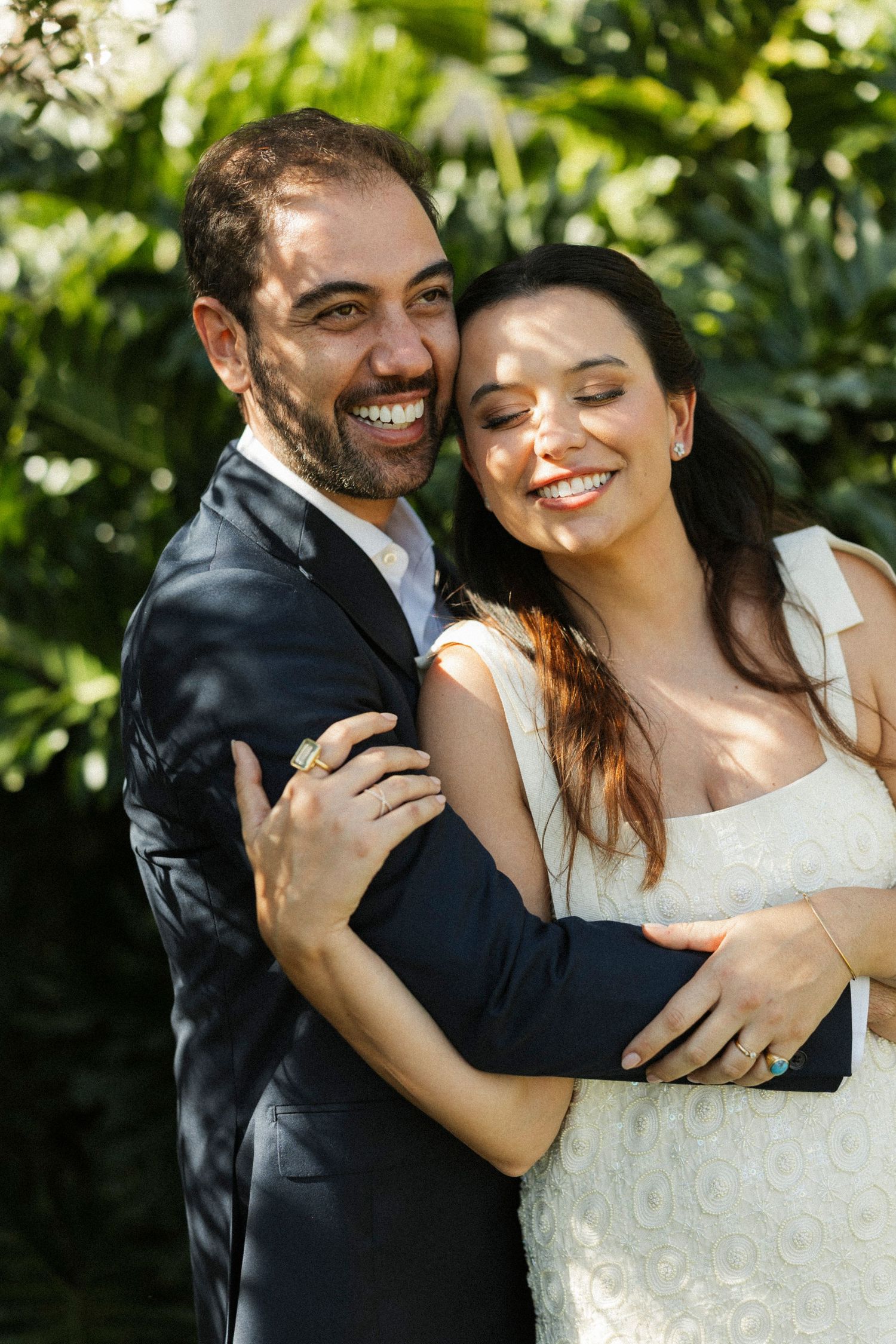 Happy couple embracing outdoors, groom in dark suit and bride in white dress, smiling among lush green foliage.