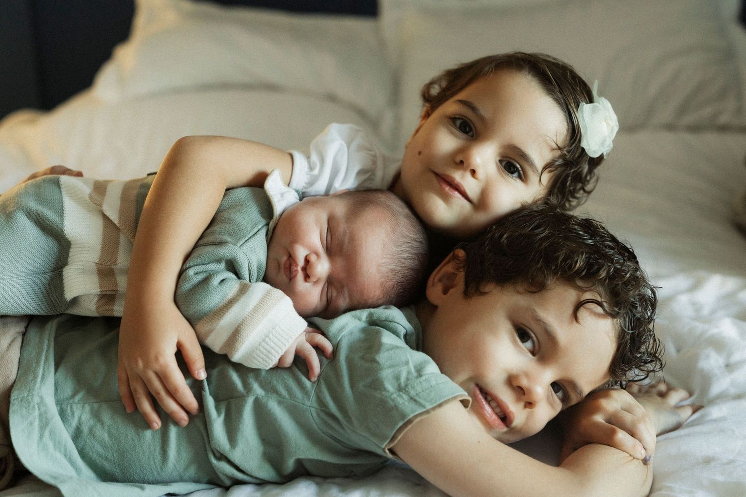 Two young siblings cuddle together with their sleeping newborn baby on a bed.