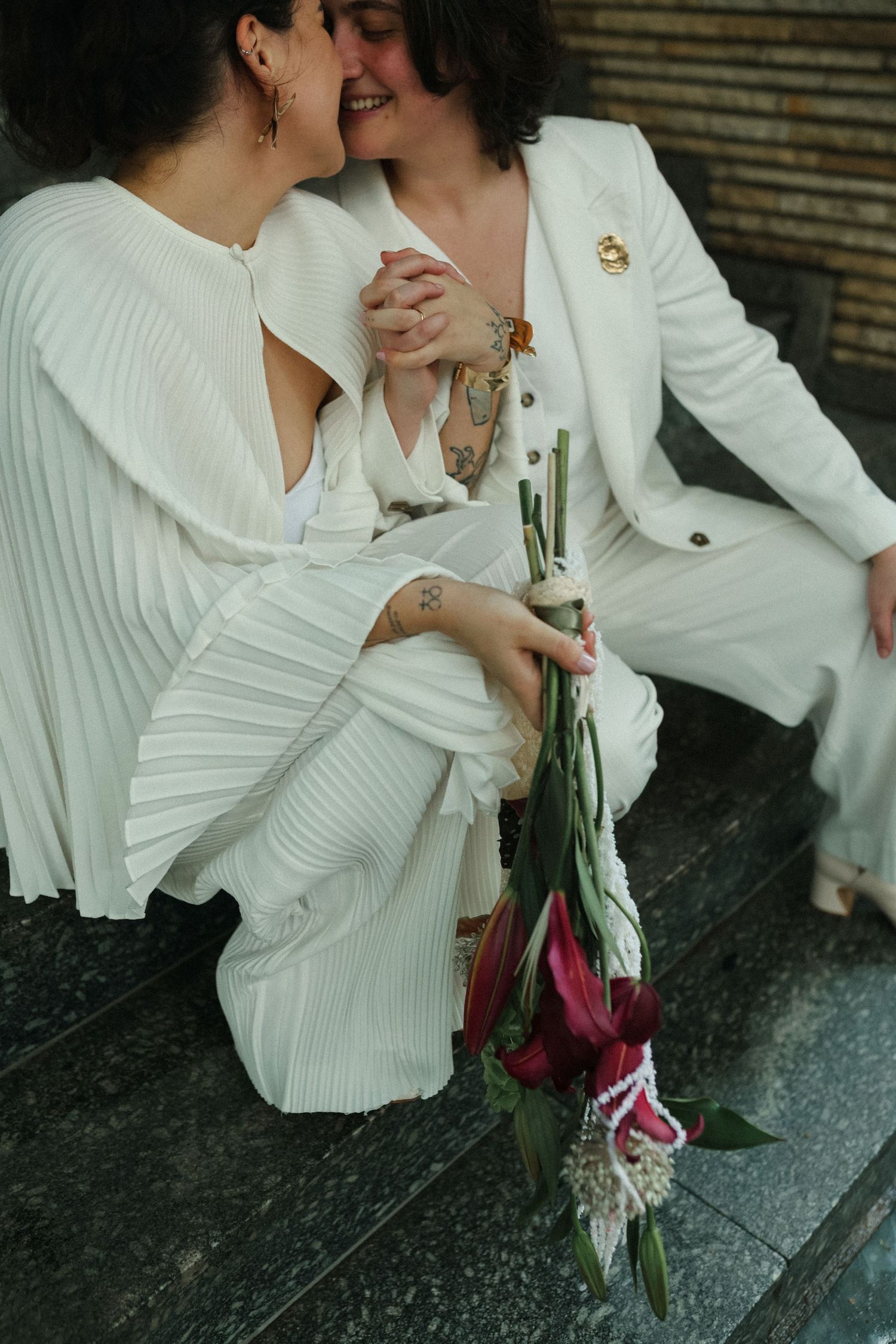 Two people in white pleated outfits share an intimate moment while holding flowers.