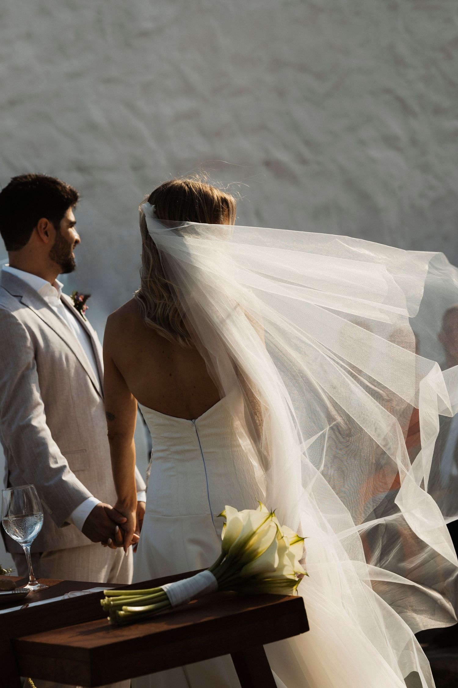 Wedding couple in white and light gray attire with flowing veil in soft lighting.