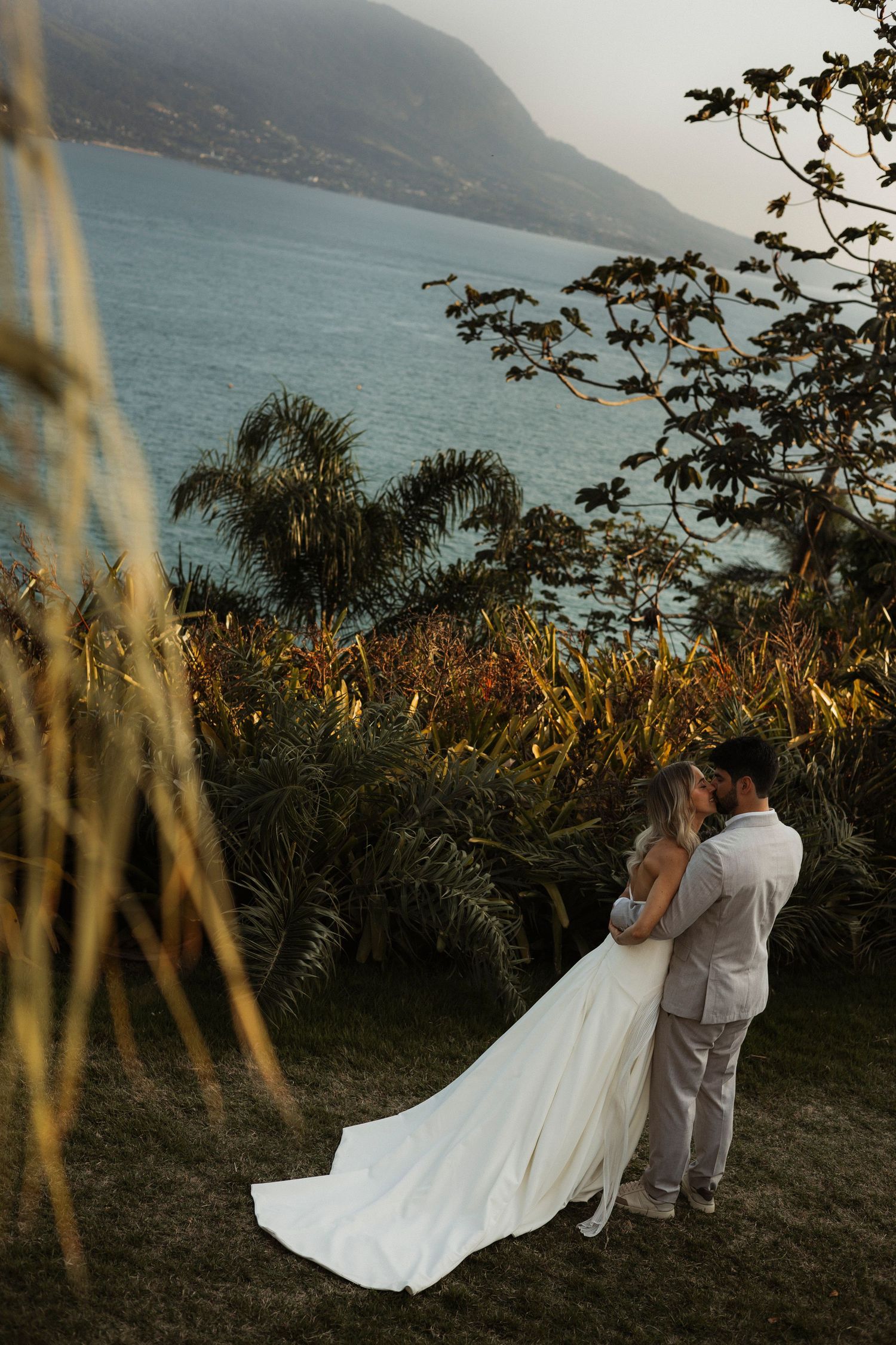 A couple in wedding attire dances on a scenic coastal hillside during golden hour.