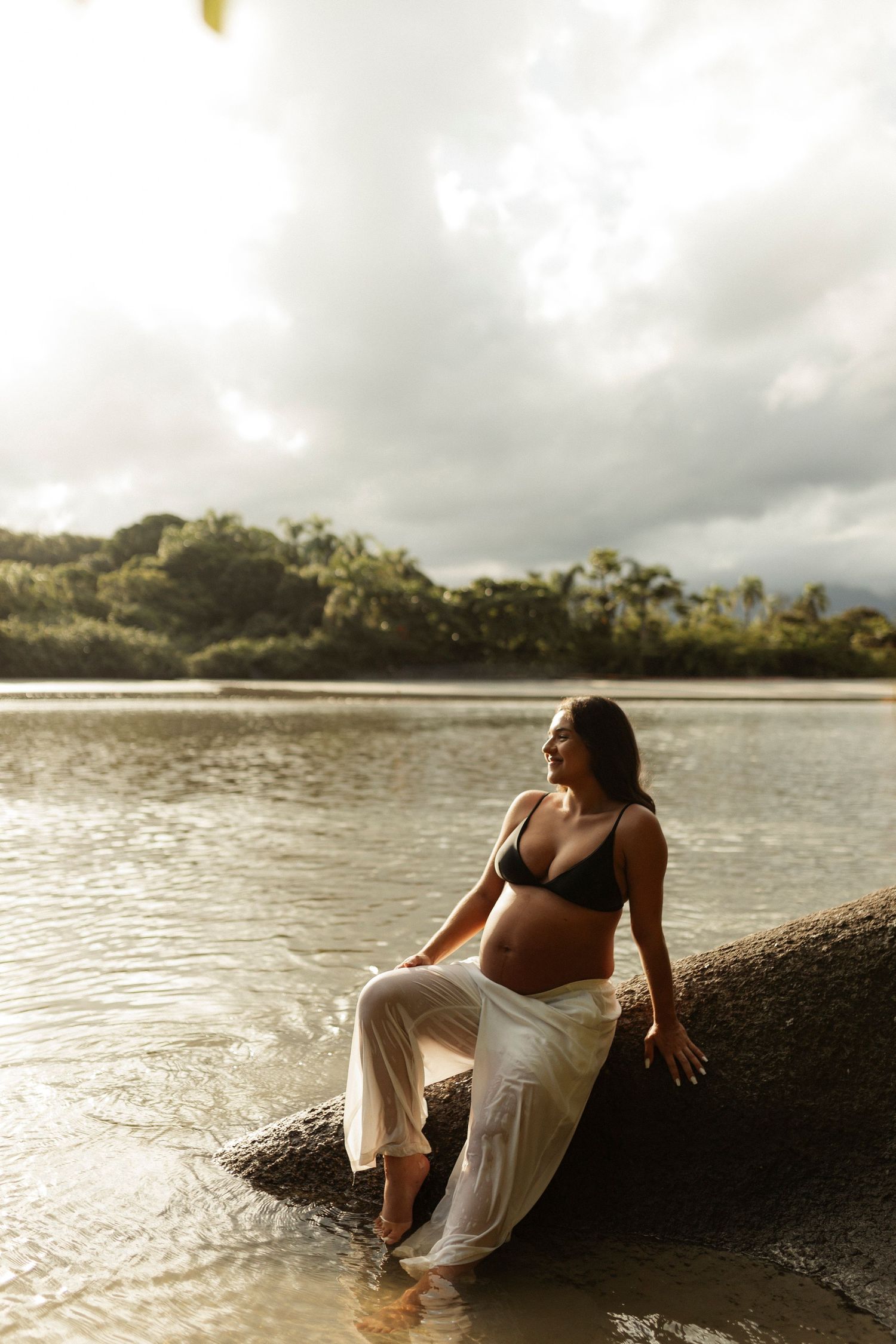 Woman in flowing white dress poses on rocky beach during golden hour.