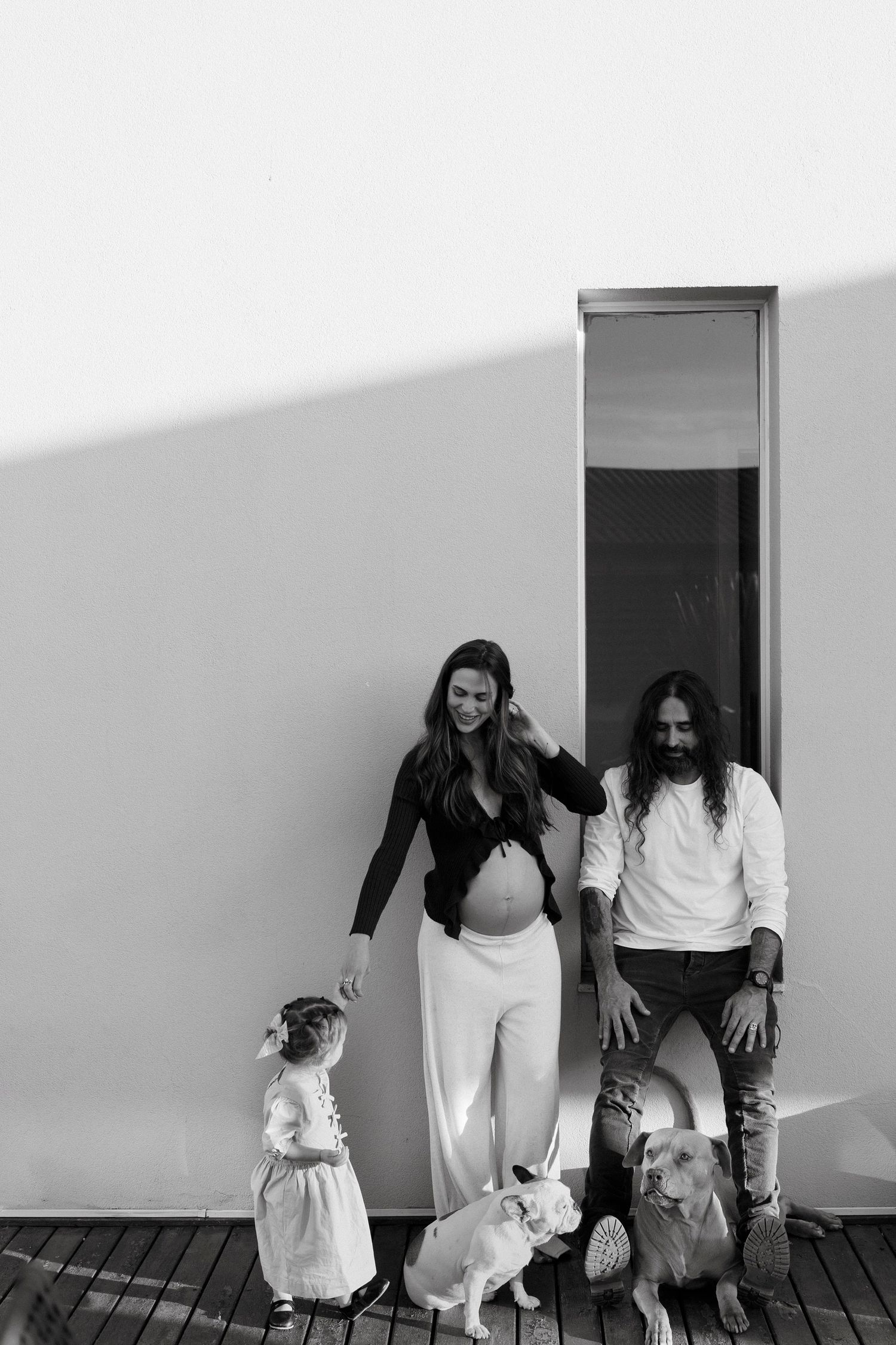 Black and white photo of a pregnant woman, man with long hair, toddler girl, and two dogs posing outside a modern white building.