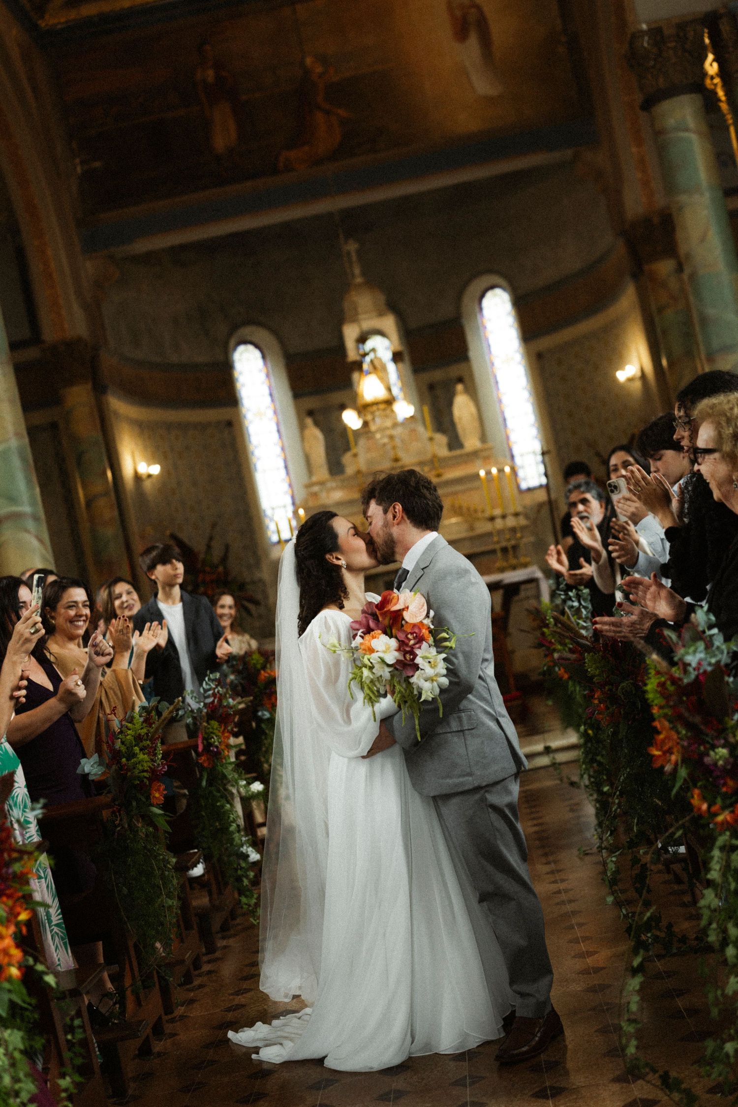Bride and groom share first kiss in ornate church aisle, surrounded by floral arrangements and applauding guests.
