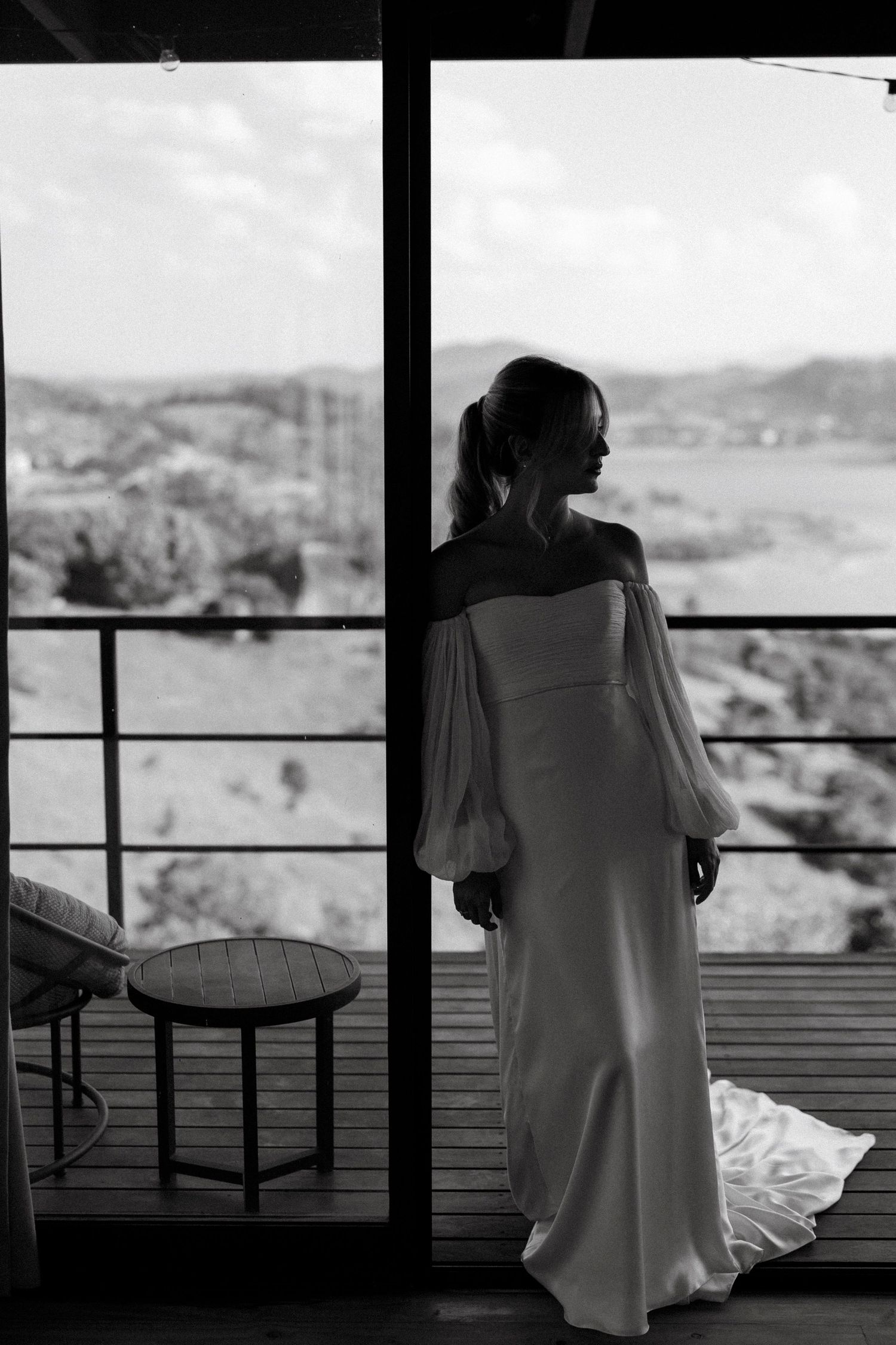 Bride in off-shoulder white gown stands by glass doors with scenic landscape view in black and white wedding photo.