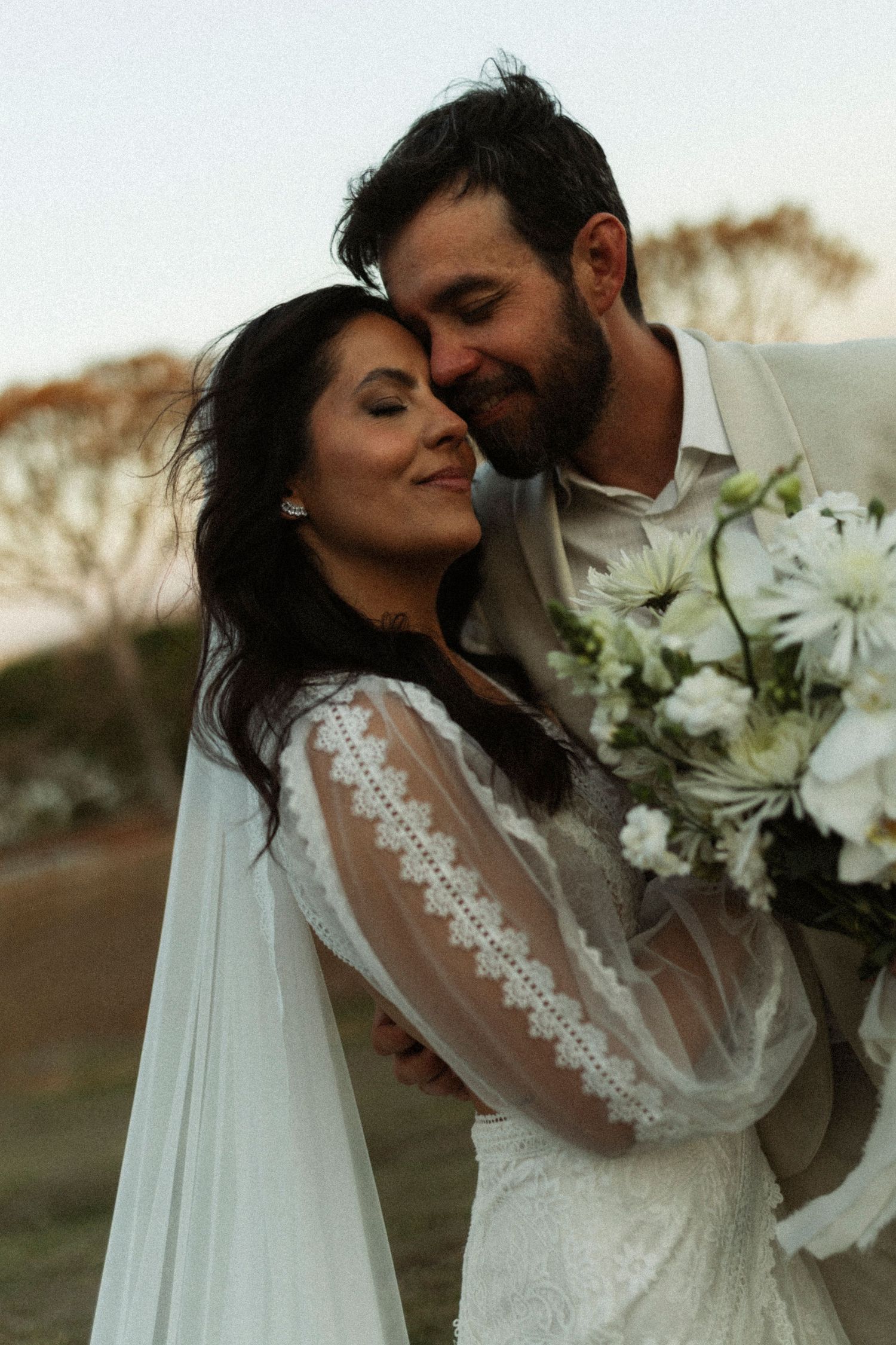 Romantic wedding couple embraces while holding white flowers during an outdoor photoshoot at sunset.
