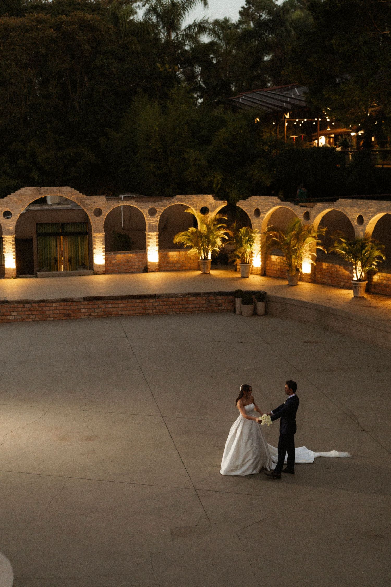 Bride and groom share a romantic moment on a grand stone plaza at dusk, framed by illuminated arches and lush palms.