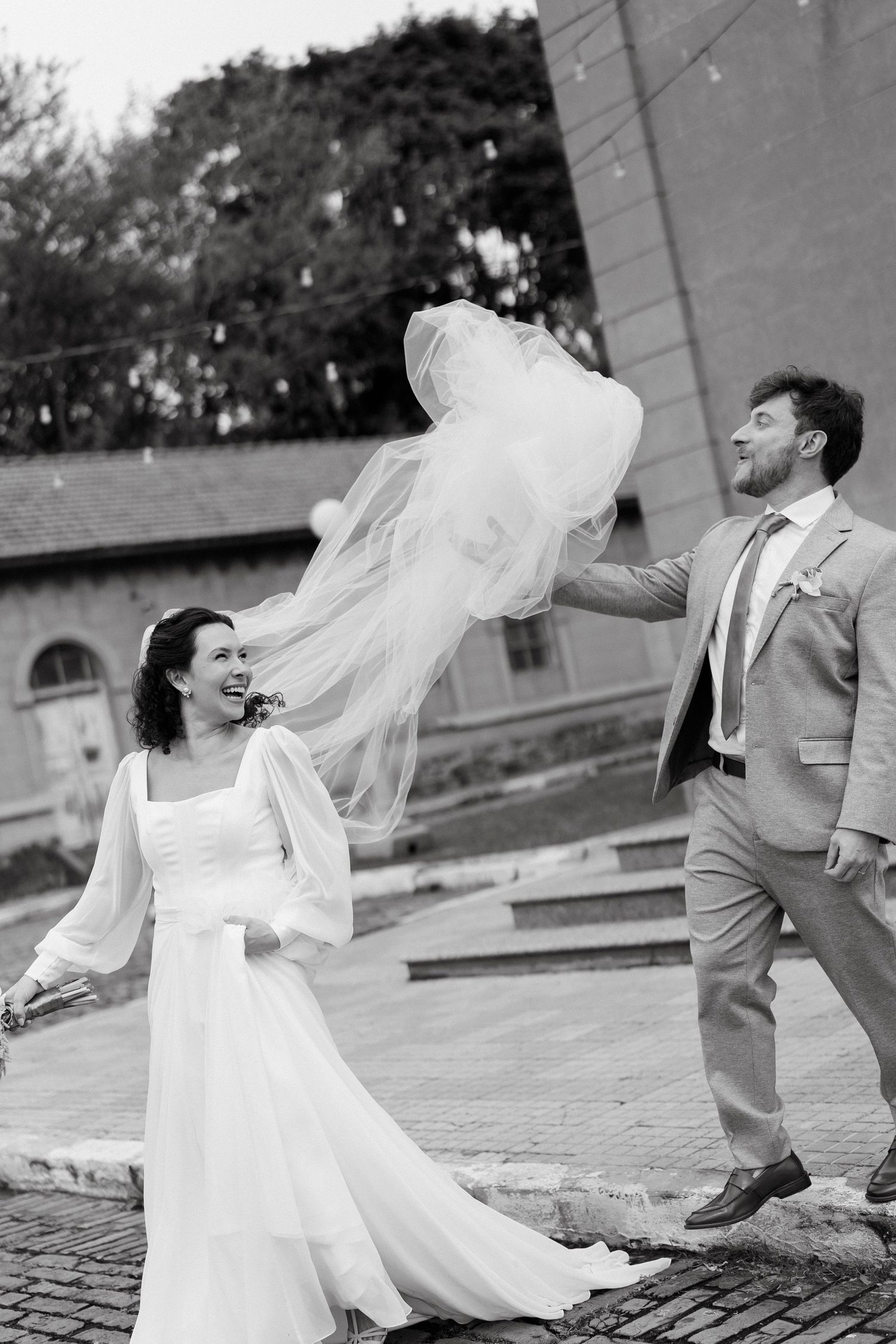 Black and white photo of a joyful bride and groom, groom tossing veil in the air outdoors on their wedding day.