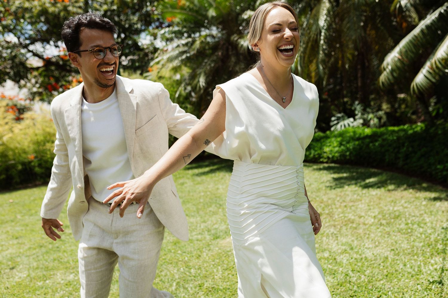 Playful couple in white outfits running and laughing together in a sunny garden setting.