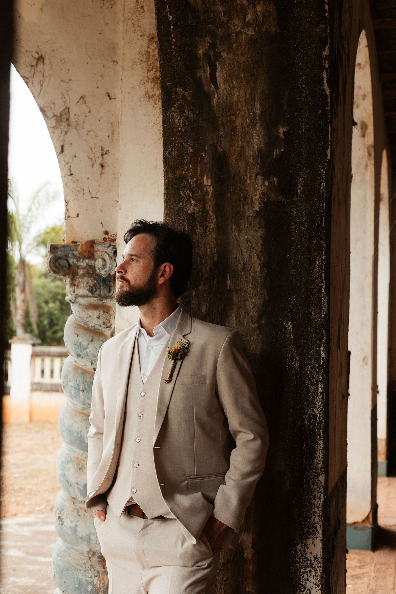 Groom in light beige suit poses thoughtfully under rustic archways.