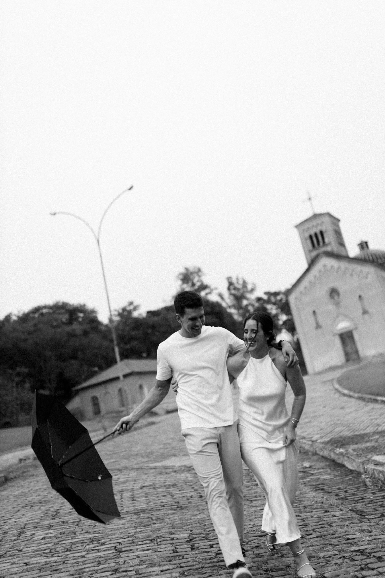 Couple in white walking along cobblestone path with umbrella and church in background.