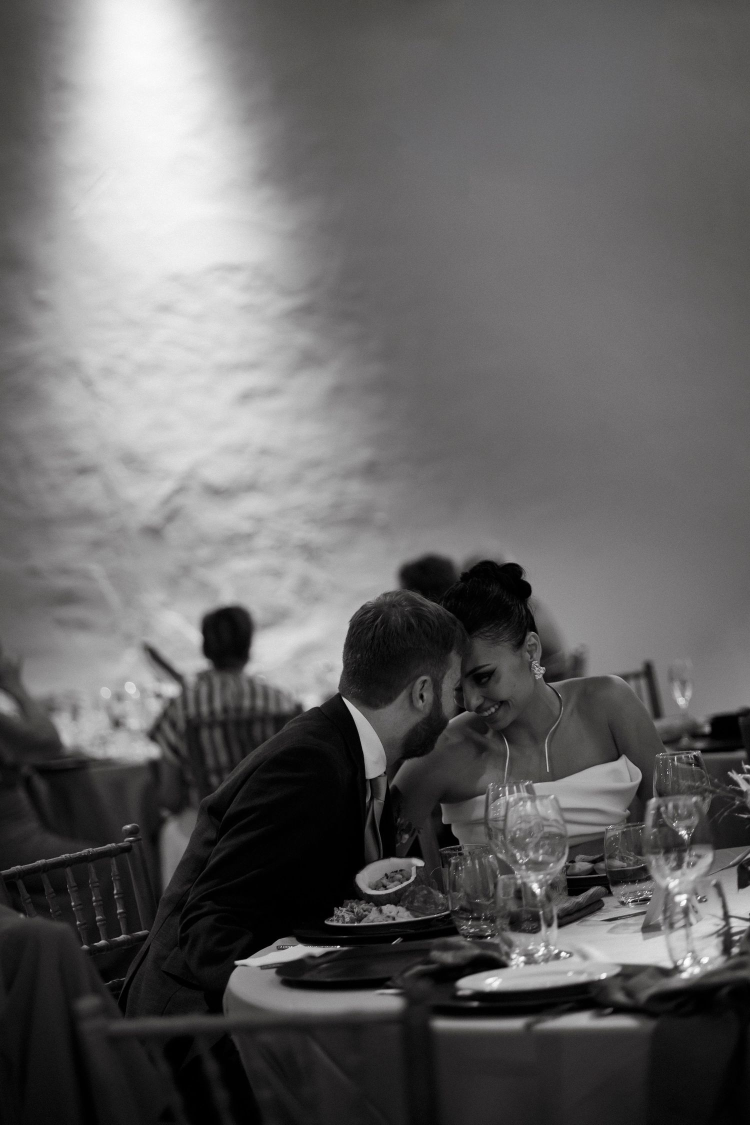 Bride and groom share an intimate moment at their wedding reception dinner table in black and white.