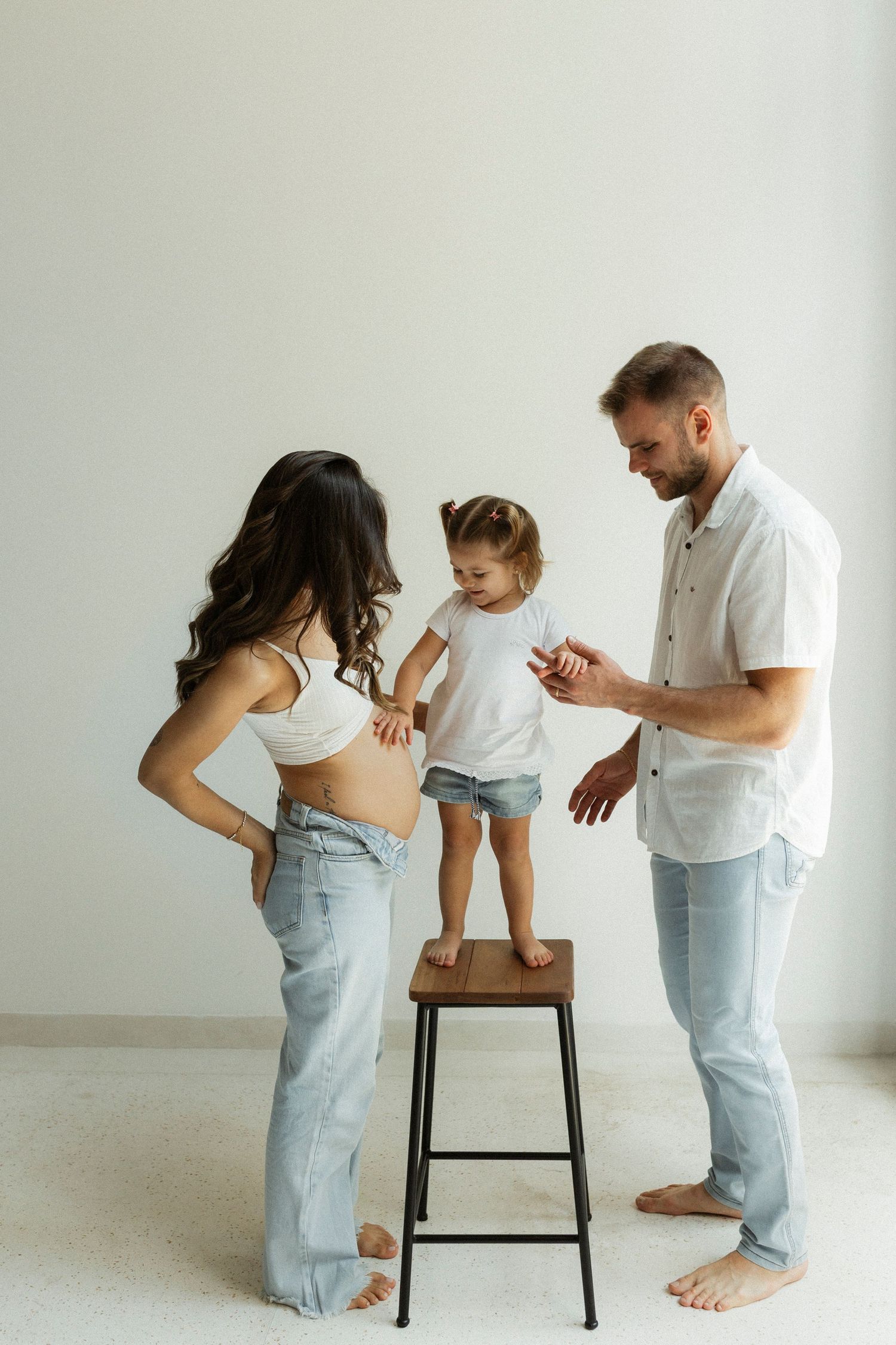 A family in casual attire stands together in a minimalist white room with a small child balanced on a wooden stool.