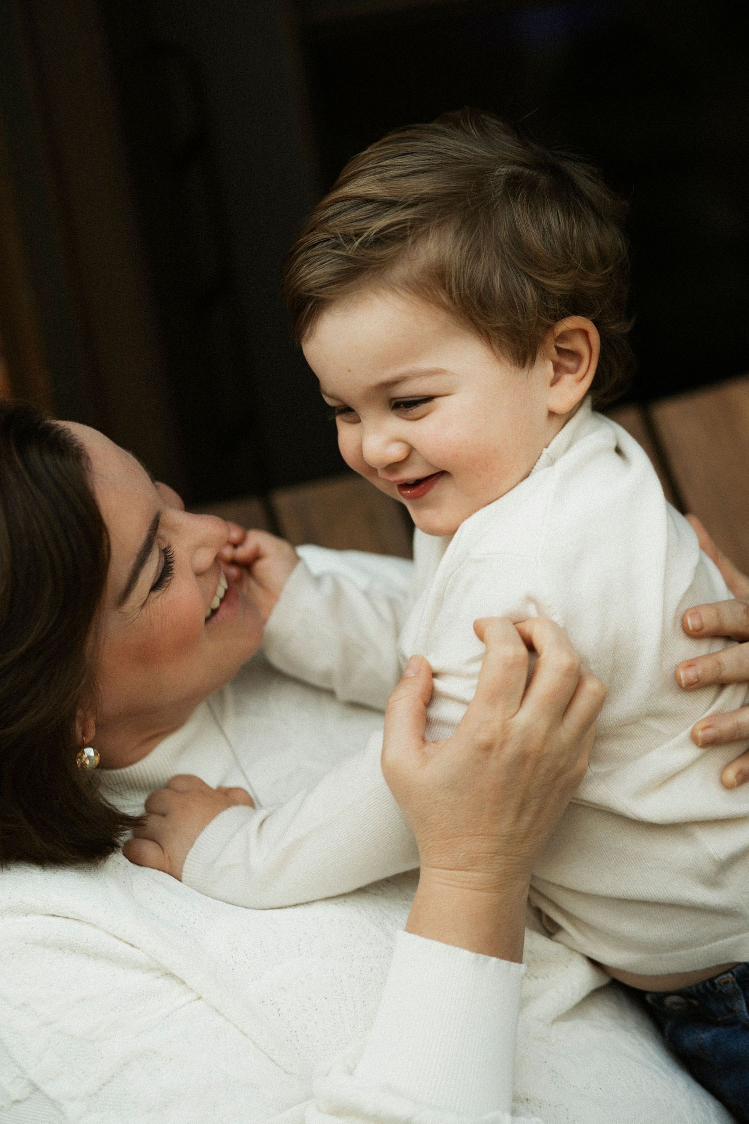 A joyful baby in white outfit laughs while being held by a smiling dark-haired mother on a white bed.