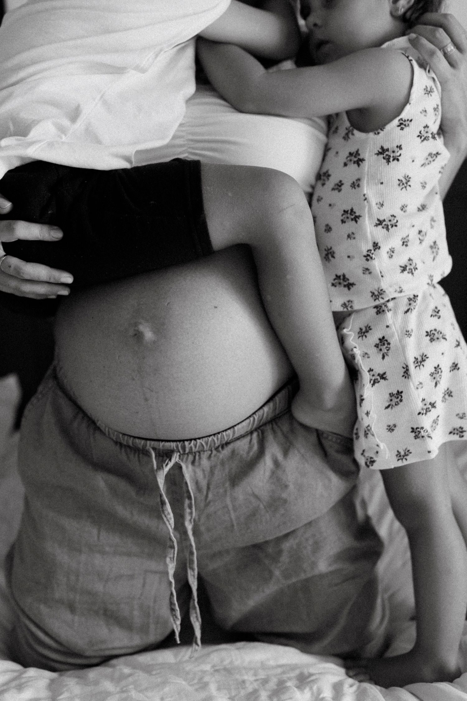 Black and white photo of pregnant woman with baby bump, embraced by partner and young child on bed.