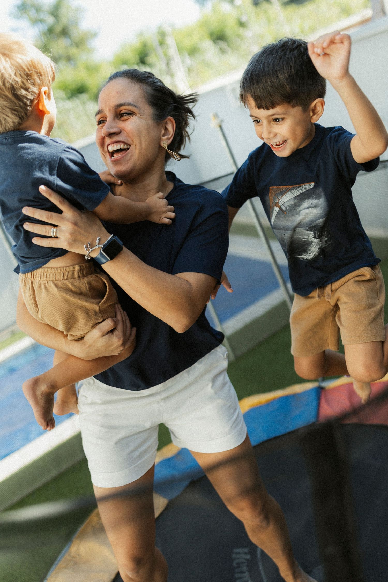 A woman laughs while holding a child on a trampoline as another boy jumps beside her outdoors.