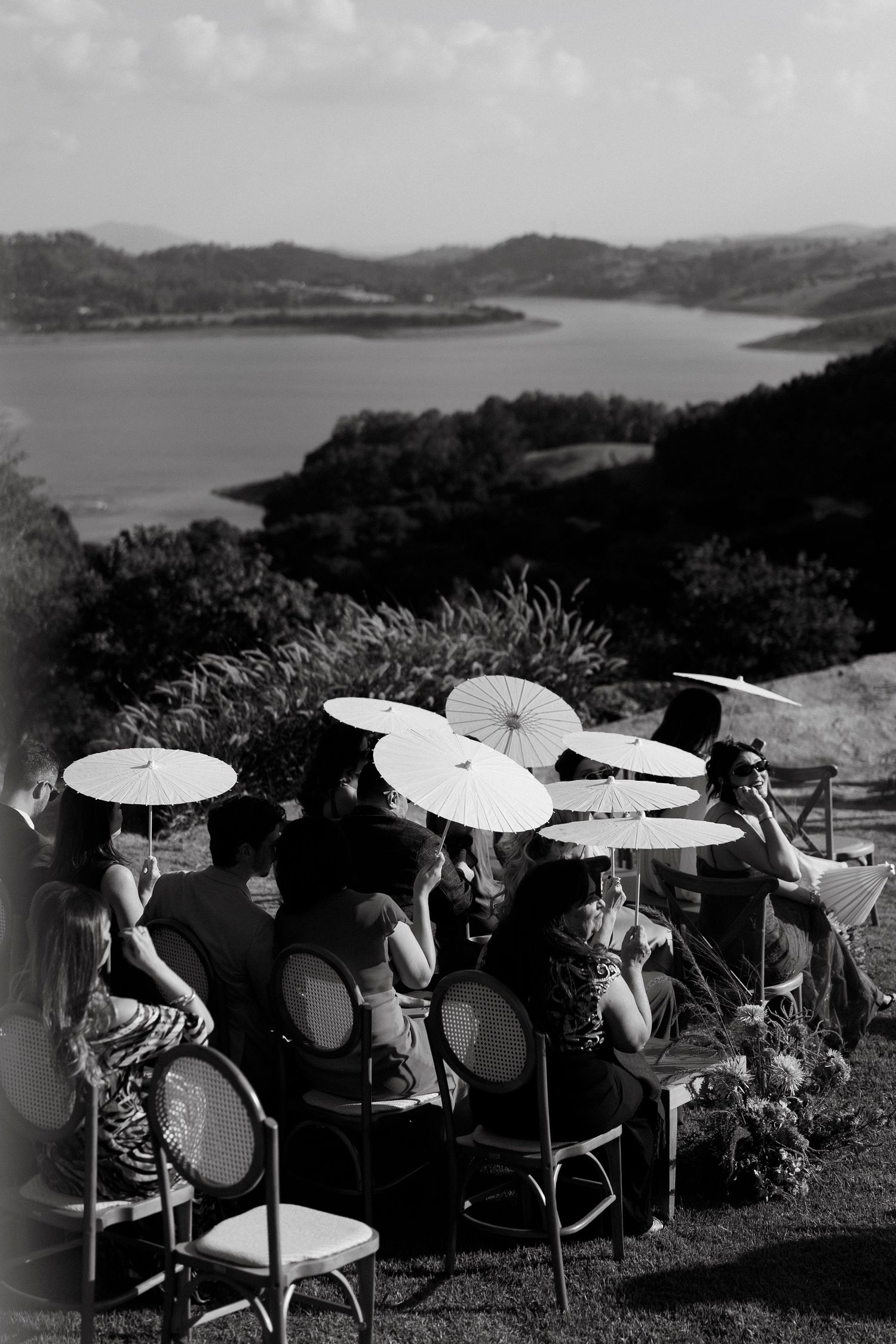 Black and white photo of wedding guests holding parasols seated outdoors with scenic lake and hills in background.