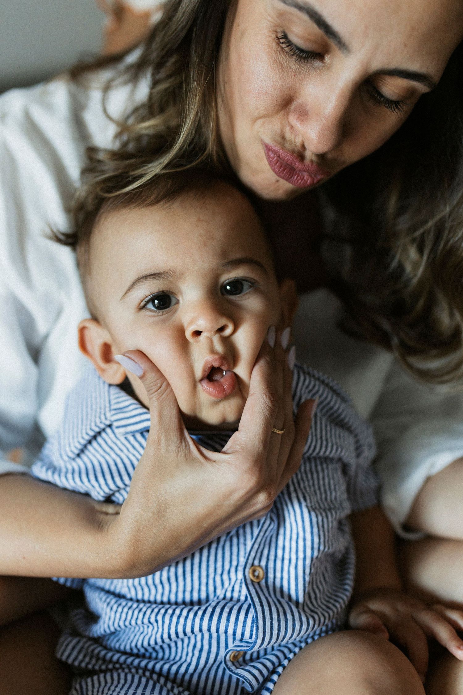 A mother tenderly holds her baby boy in a blue striped outfit, leaning in for a kiss while he looks at the camera.
