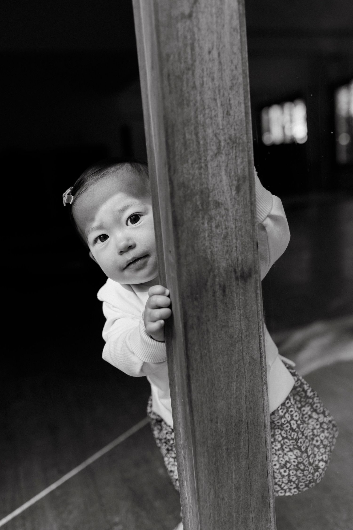 Black and white photo of a curious baby peeking around a wooden post, holding on with tiny hands.