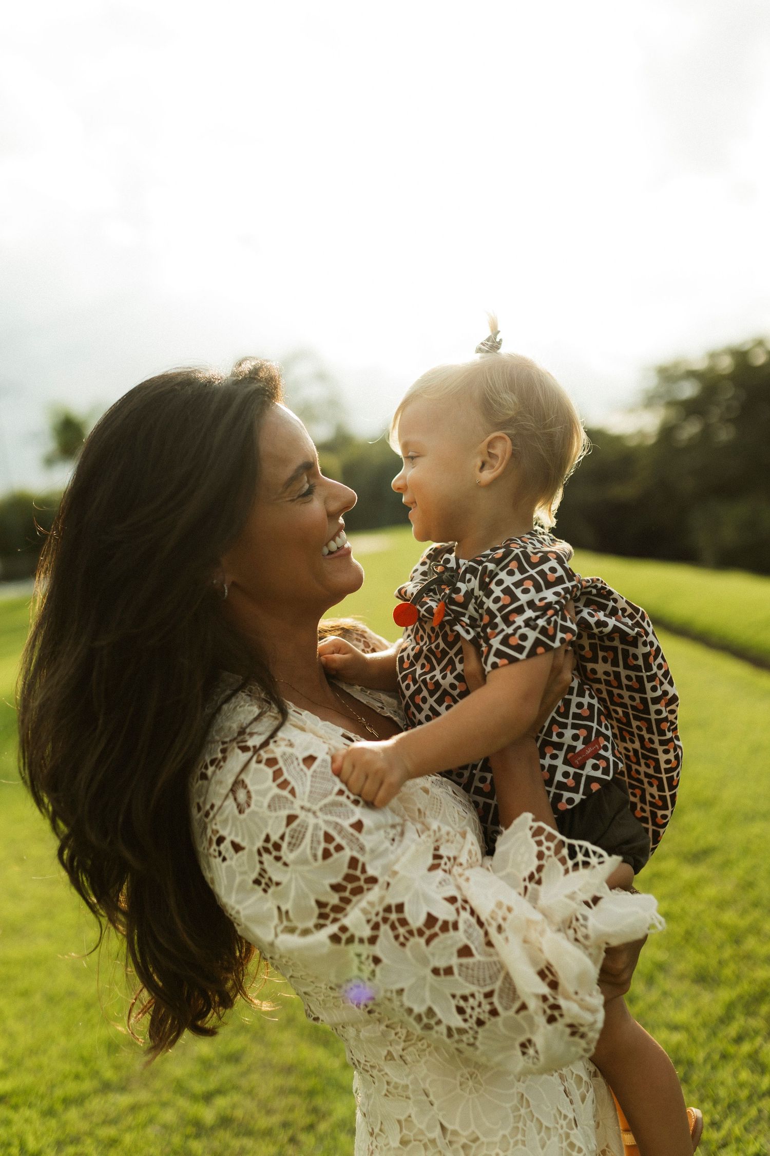 A smiling mother in white lace dress holds her baby outdoors in golden sunlight on a green lawn.