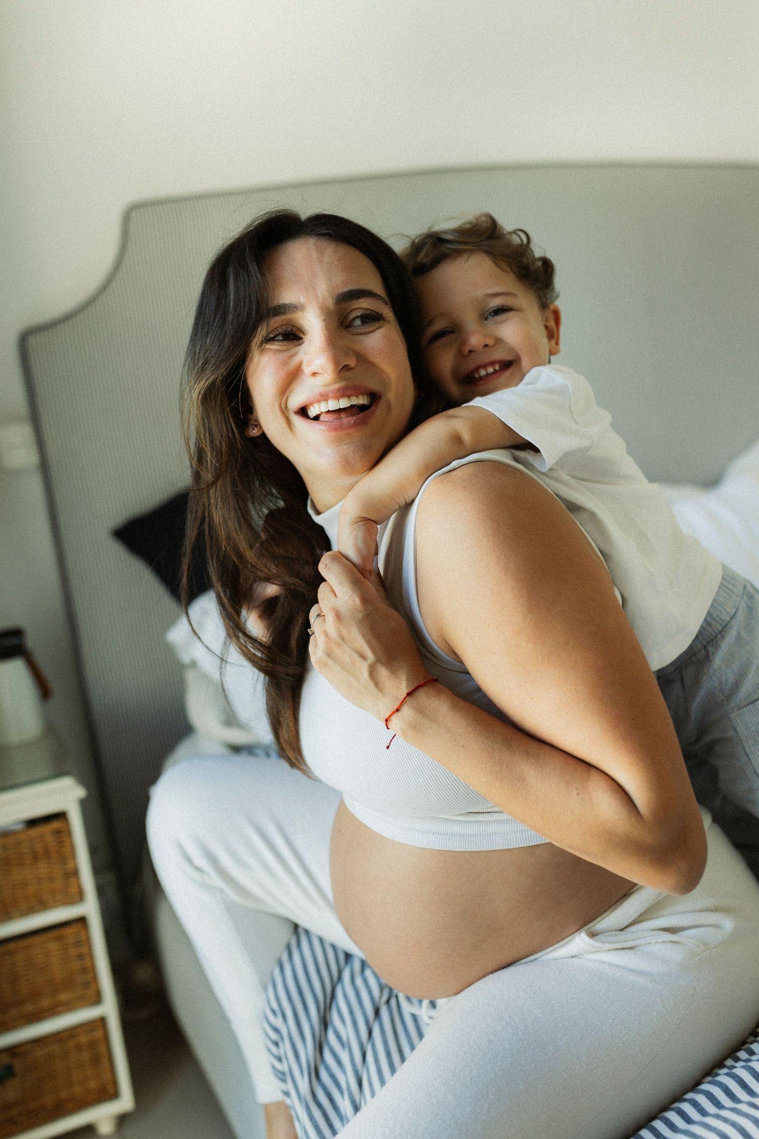 Smiling pregnant woman in white crop top holding a happy toddler on her back while sitting on a bed.