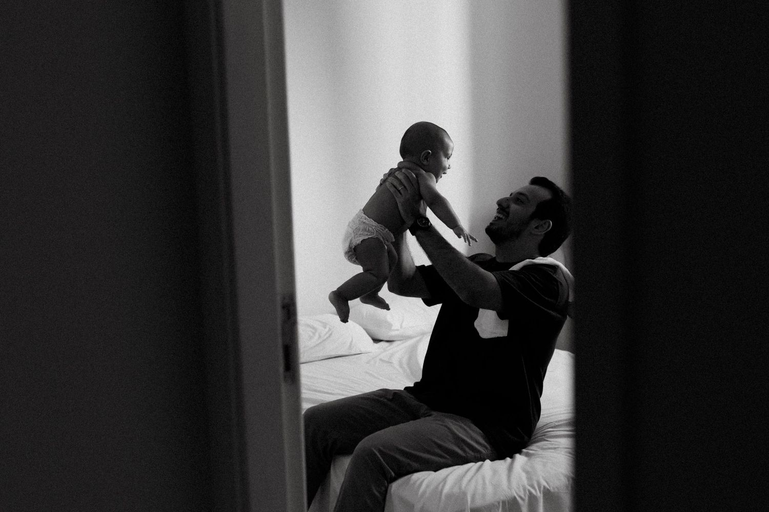 Black and white photo of a father lifting his baby joyfully while sitting on a bed, viewed through a doorway.