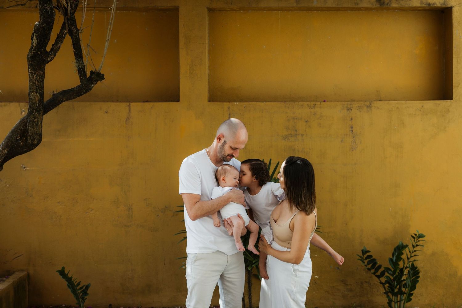 A couple in white outfits lovingly kisses their newborn baby against a warm yellow wall outdoors.