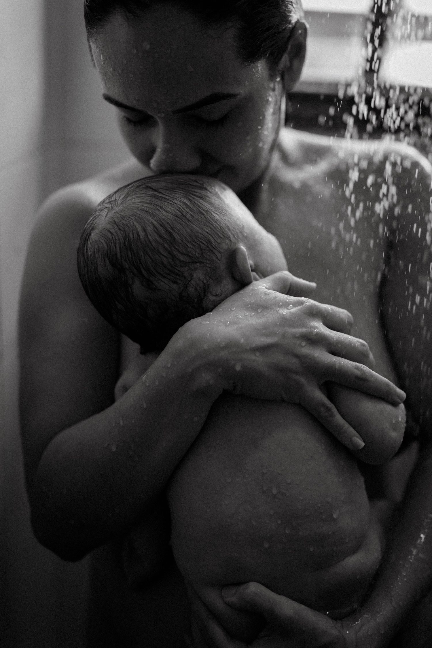 Black and white photo of a mother tenderly kissing her newborn baby while bathing together in a shower.
