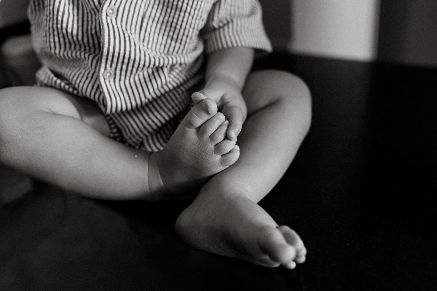Black and white photo of a baby sitting cross-legged, showing chubby feet and hands in a striped outfit.