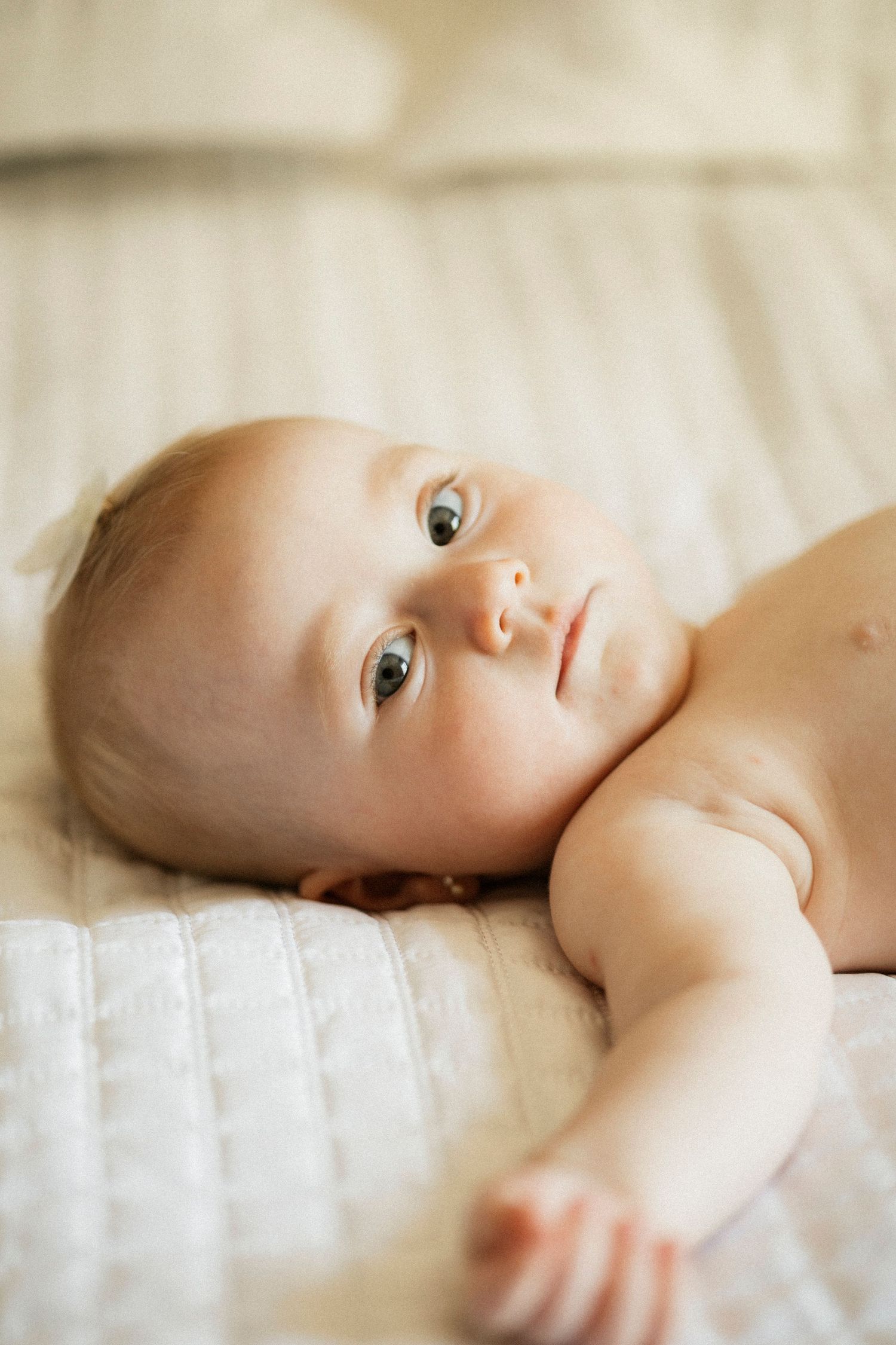 A peaceful newborn baby with blue eyes lying on a white quilted blanket, gazing upward with a calm expression.